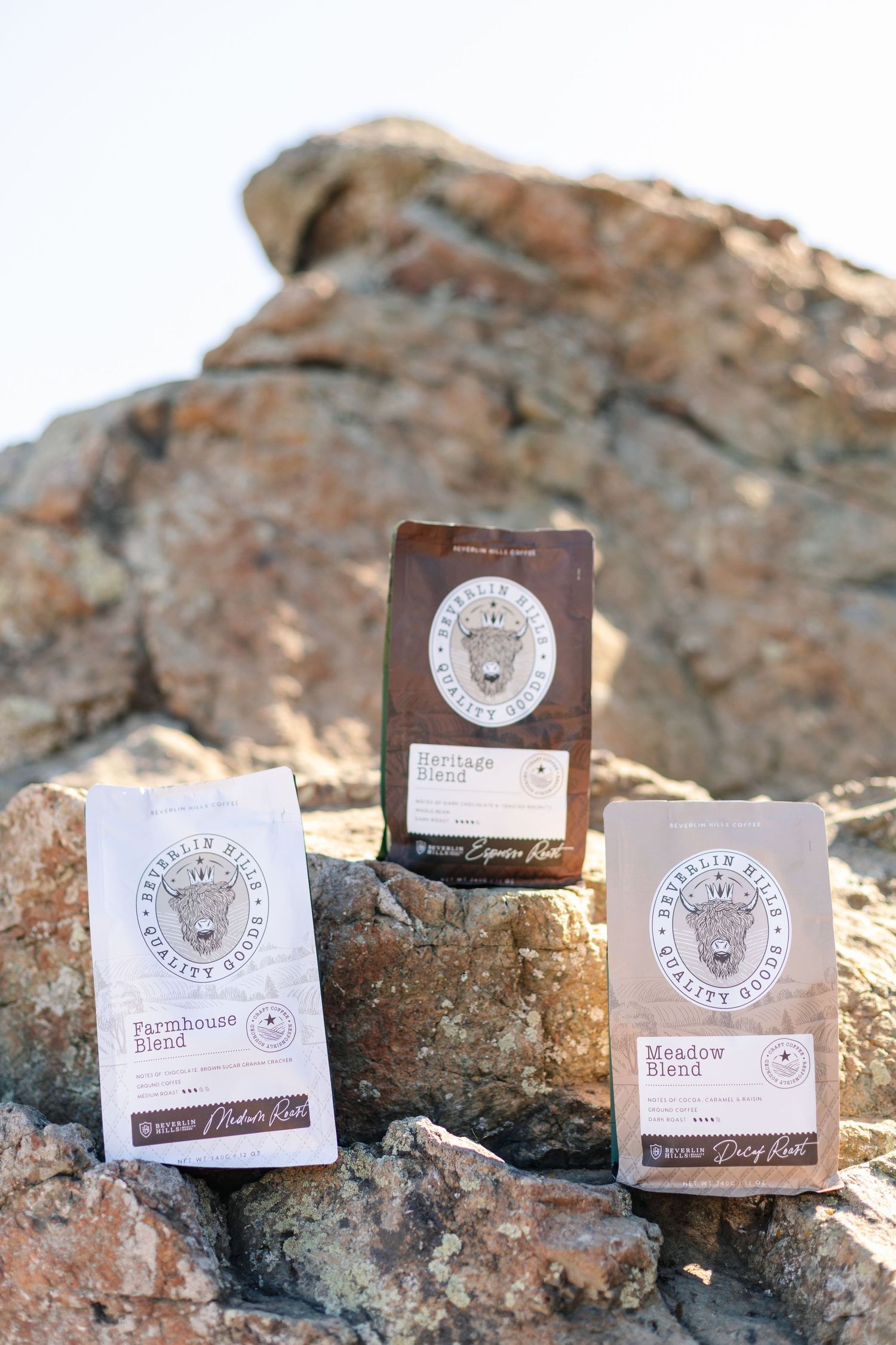 Three Beverlin Hills Quality Goods coffee blend bags arranged on rocky outdoor terrain against sky.