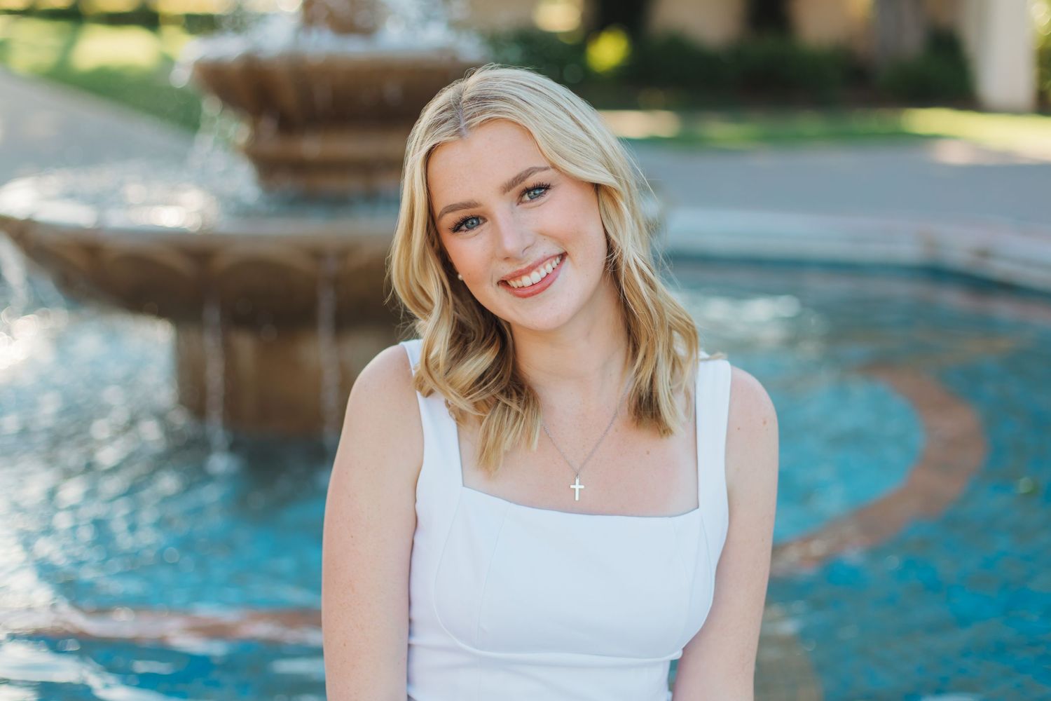 A smiling blonde young woman in a white dress sits relaxed near a sparkling fountain.