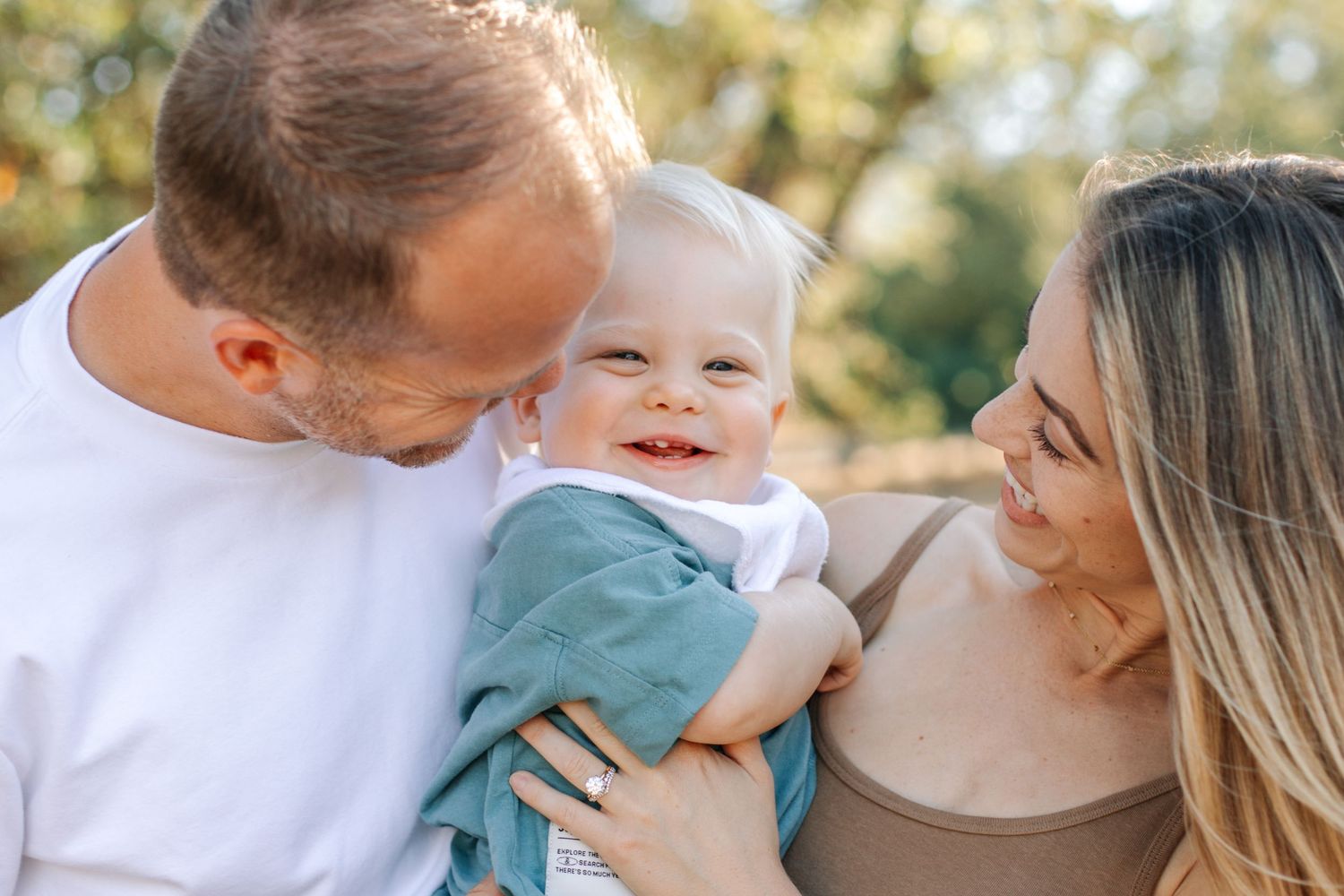 Two parents kissing and cuddling their smiling baby boy outdoors in warm sunlight.