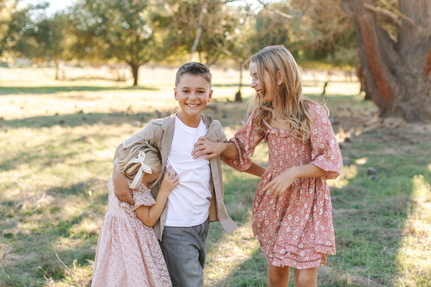 Three children laugh and play together in a sunny park with trees in the background.