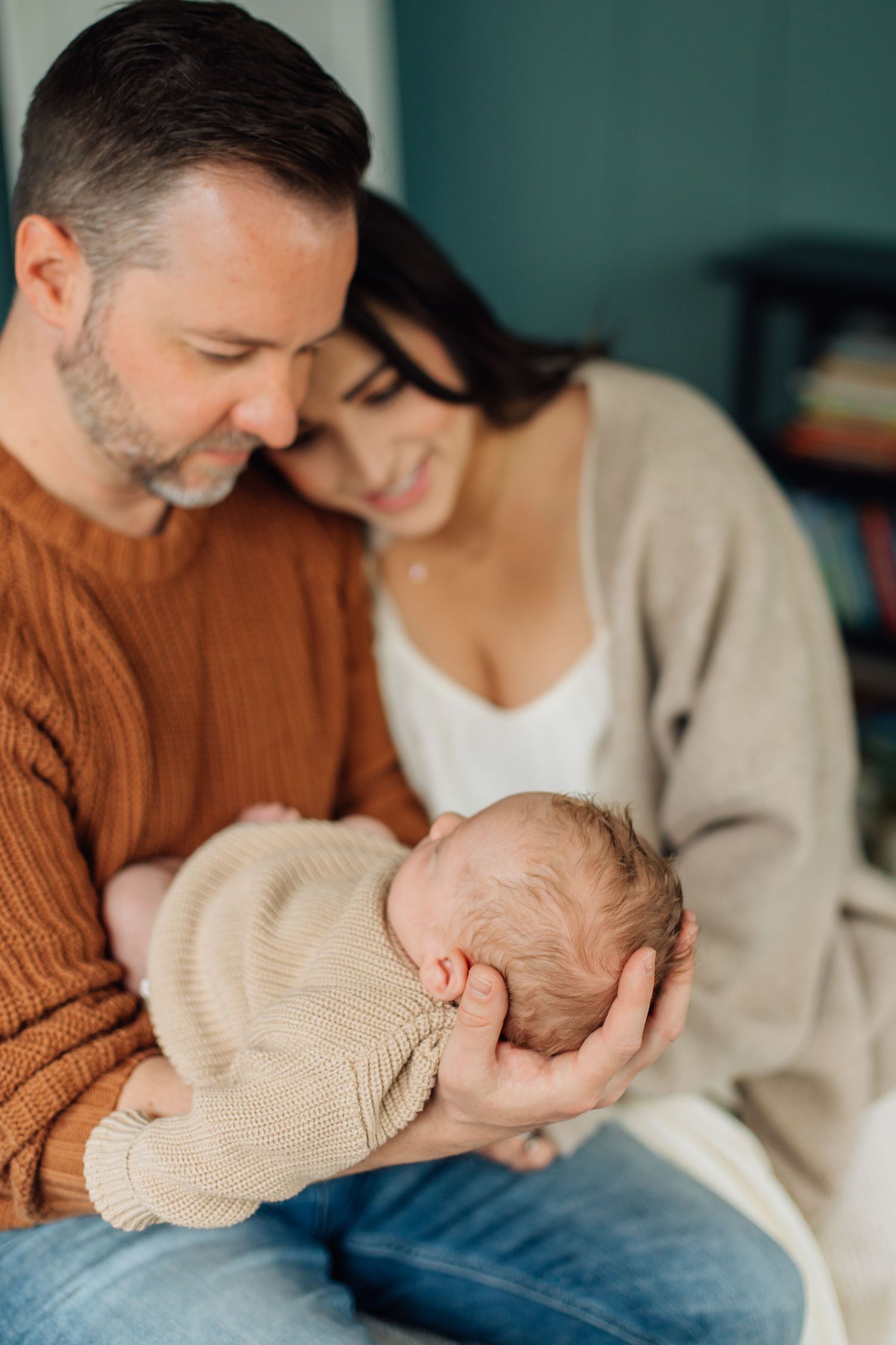 Smiling parents in cozy sweaters lovingly holding their sleeping newborn baby together.