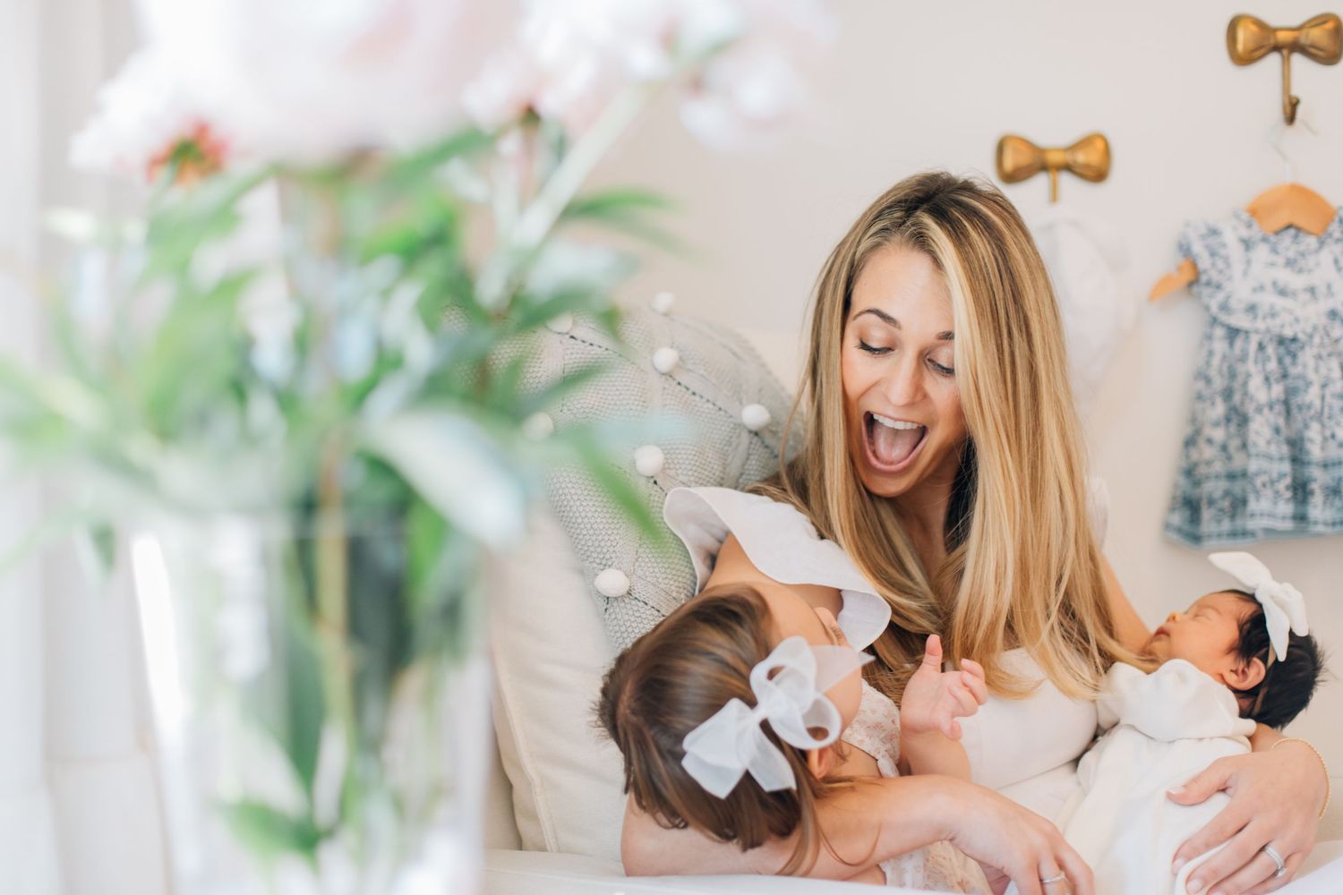 Excited mother laughs with open mouth while holding newborn and toddler daughter in bright nursery.