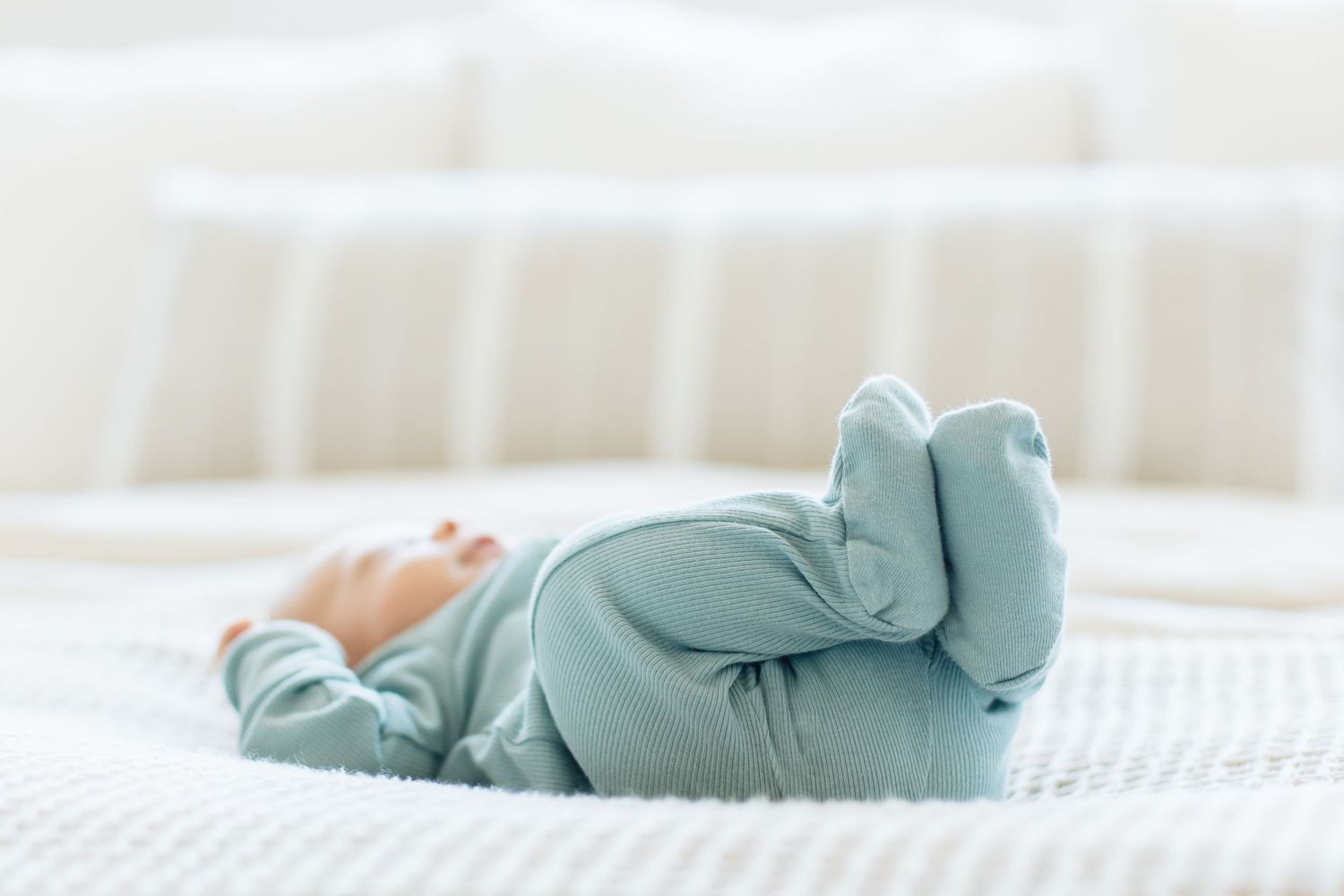 Newborn baby in light blue footed sleeper lying on back with legs raised on white textured blanket.
