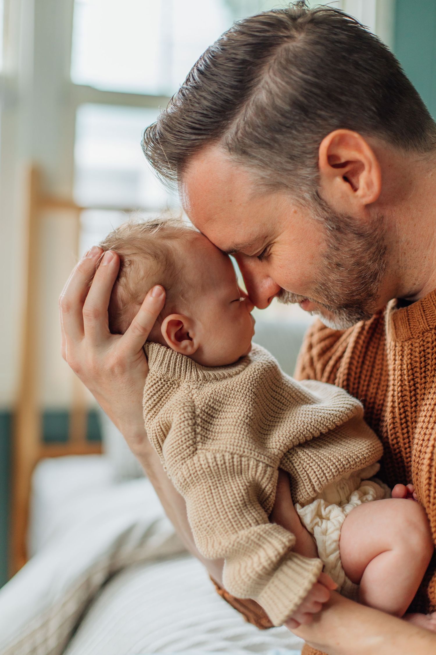 Bearded father in rust sweater touching foreheads with his newborn baby lovingly.