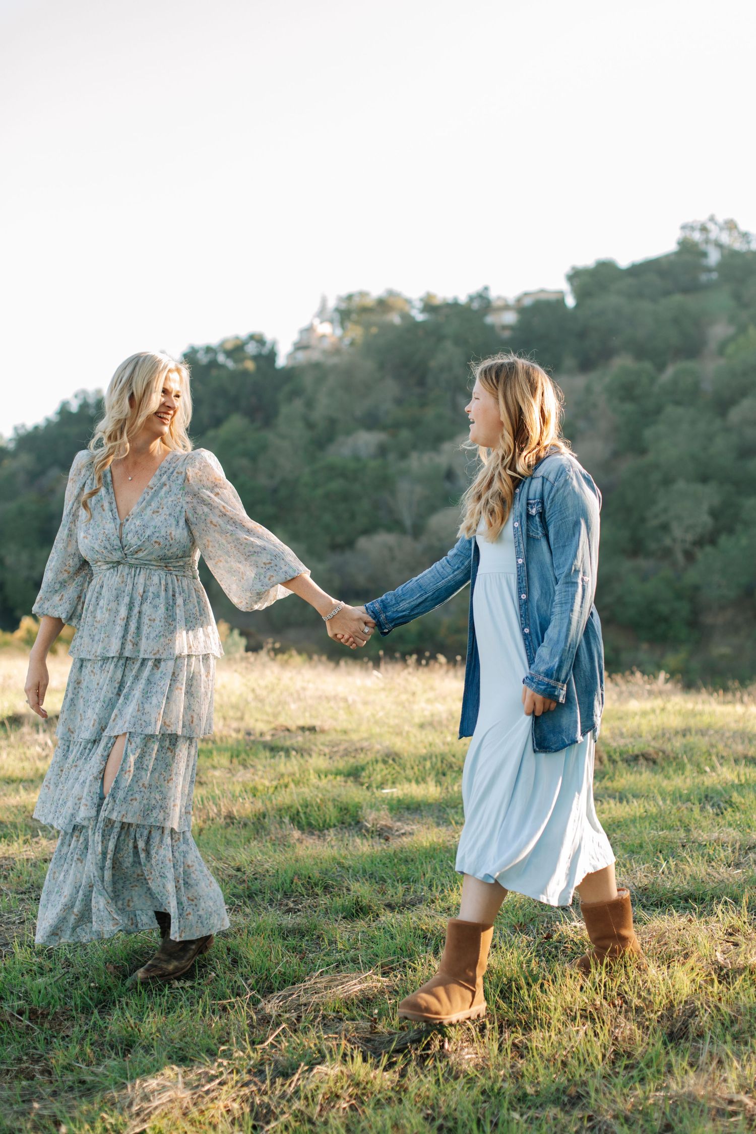 Two blonde women in bohemian dresses holding hands and walking together in a golden meadow.