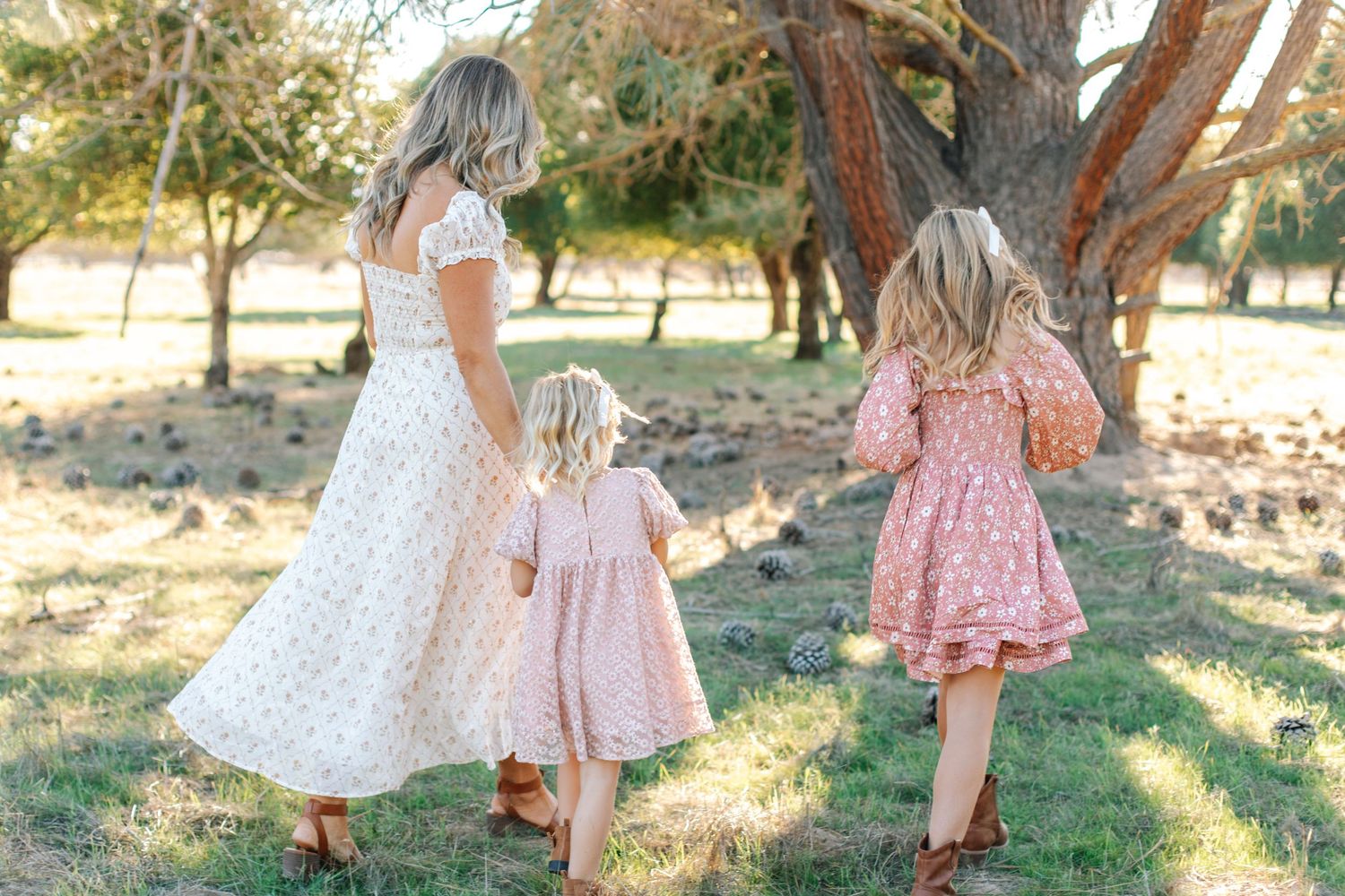 A mother and her two daughters in matching pink dresses walk hand in hand through a sunny park.