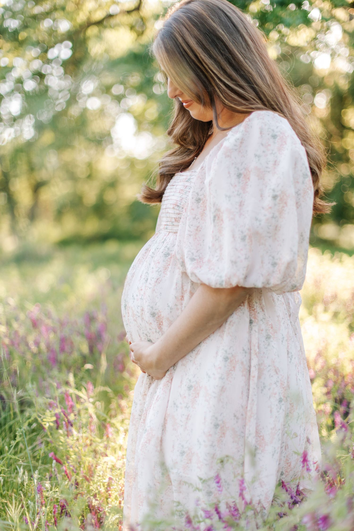 Elegant pregnant woman in floral dress cradling her baby bump in a sunny wildflower meadow.