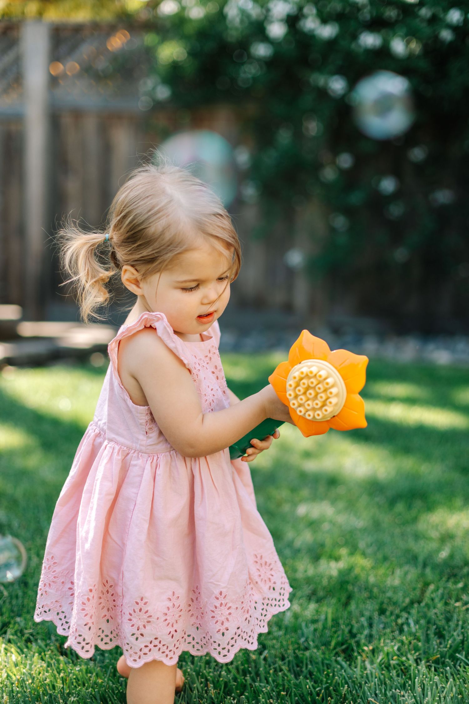 A toddler girl in a pink dress examines a sunflower-shaped bubble toy outdoors.