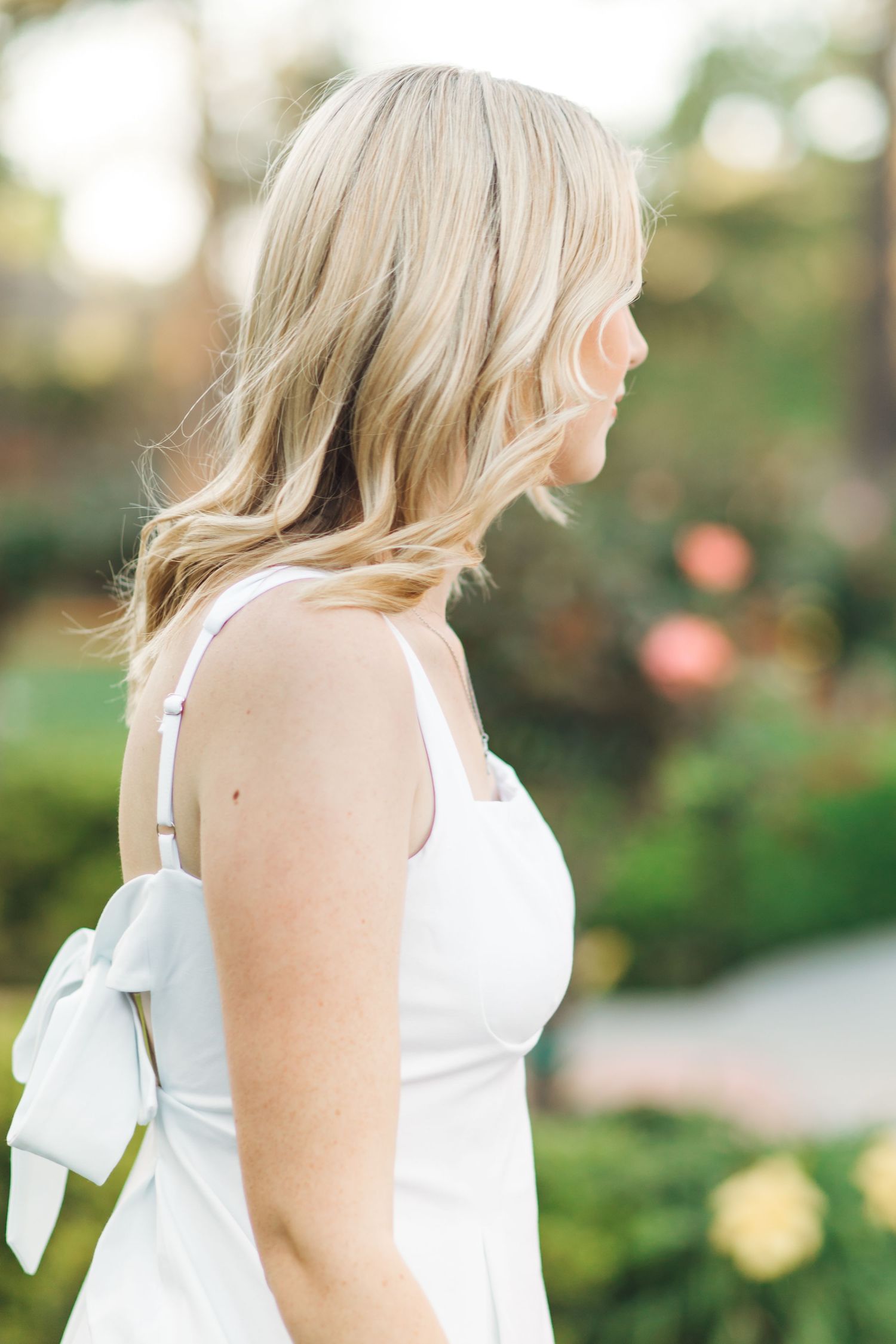 Close-up side profile of blonde woman in white bow-back dress outdoors in garden setting.
