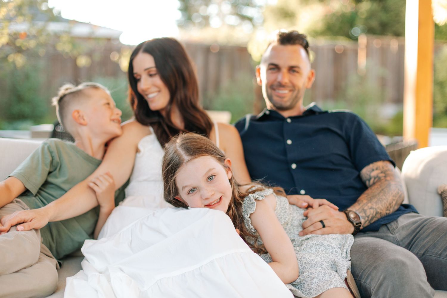 Tattooed dad and brunette mom smile with two kids on a cozy outdoor sofa in backyard.