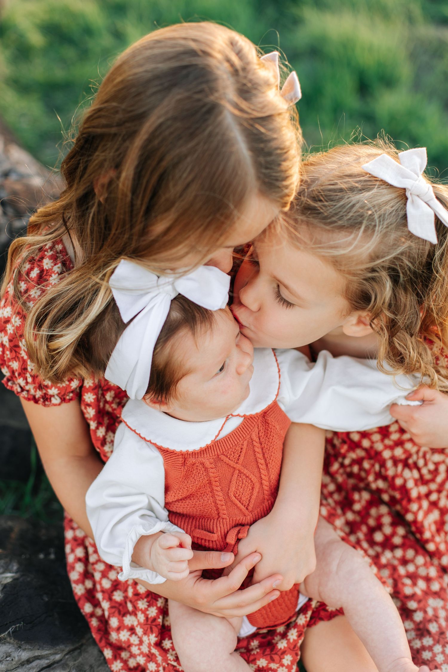 Two sisters in matching red floral dresses tenderly kiss a newborn baby sibling outdoors.