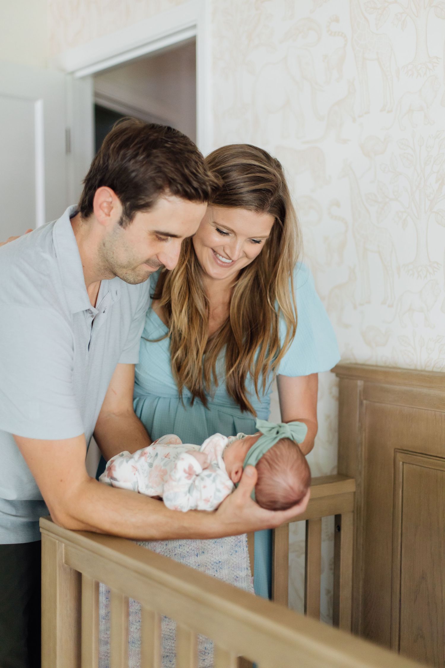 New parents smiling and admiring their swaddled newborn baby over a wooden nursery crib.