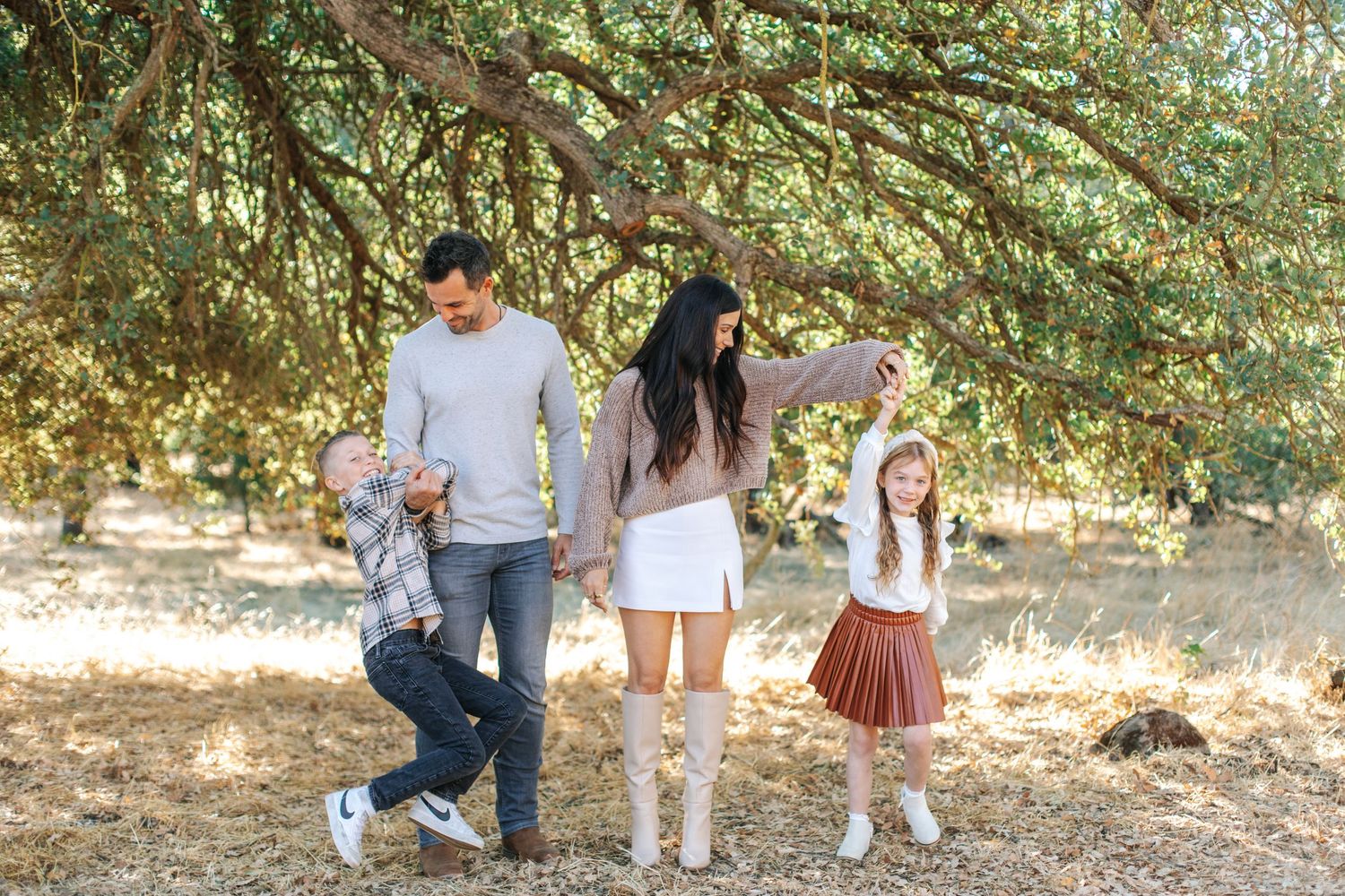 Family of four playing outdoors under a large tree, parents dancing with two young children.