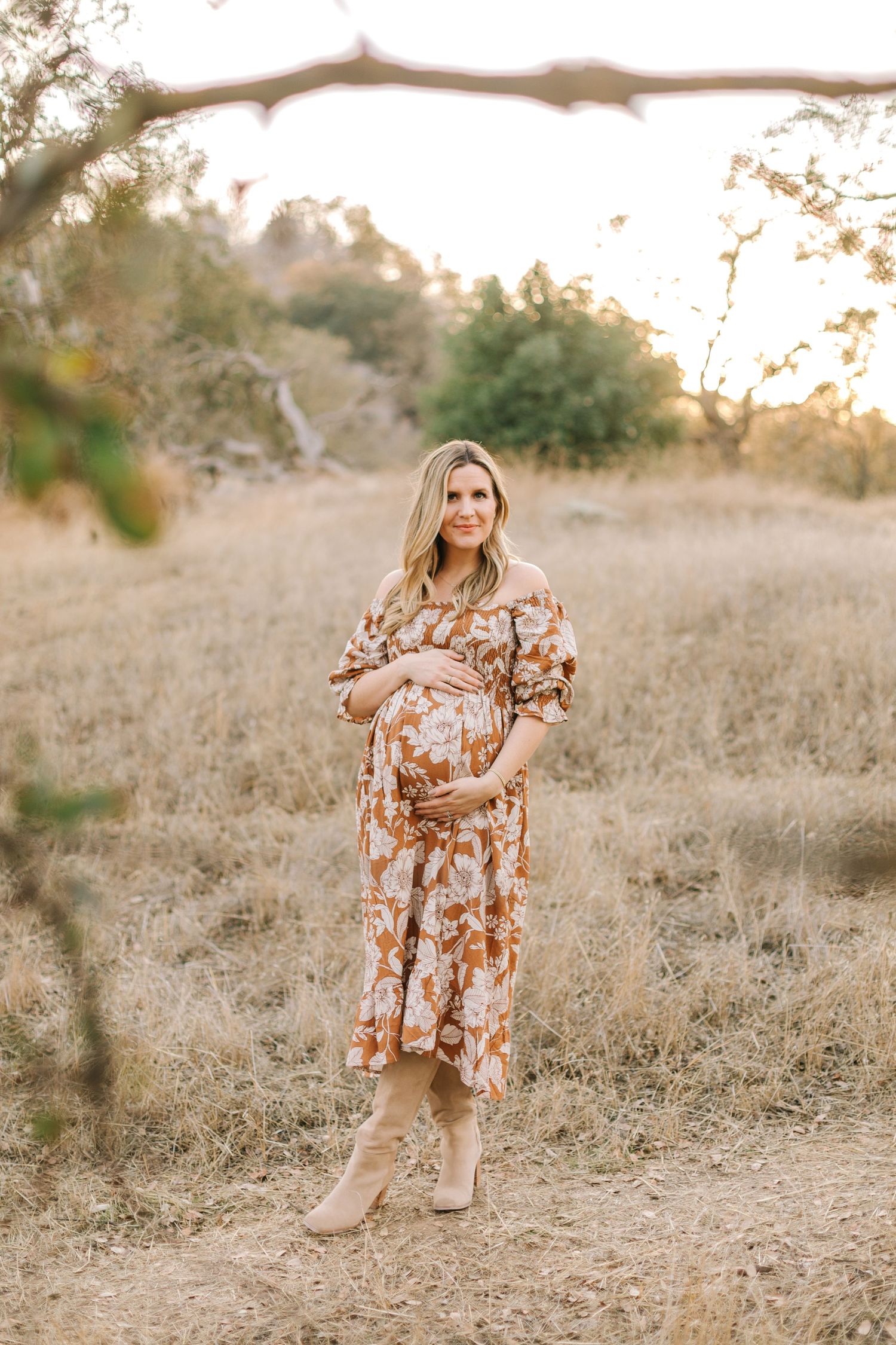 Pregnant woman in floral off-shoulder dress holding baby bump standing in a sunlit autumn field.