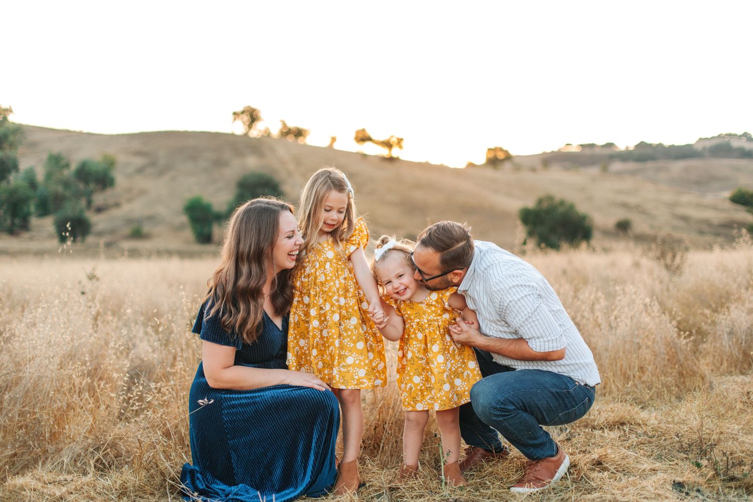 Family of four sharing a joyful moment in a golden field, parents kissing toddler in yellow dress.