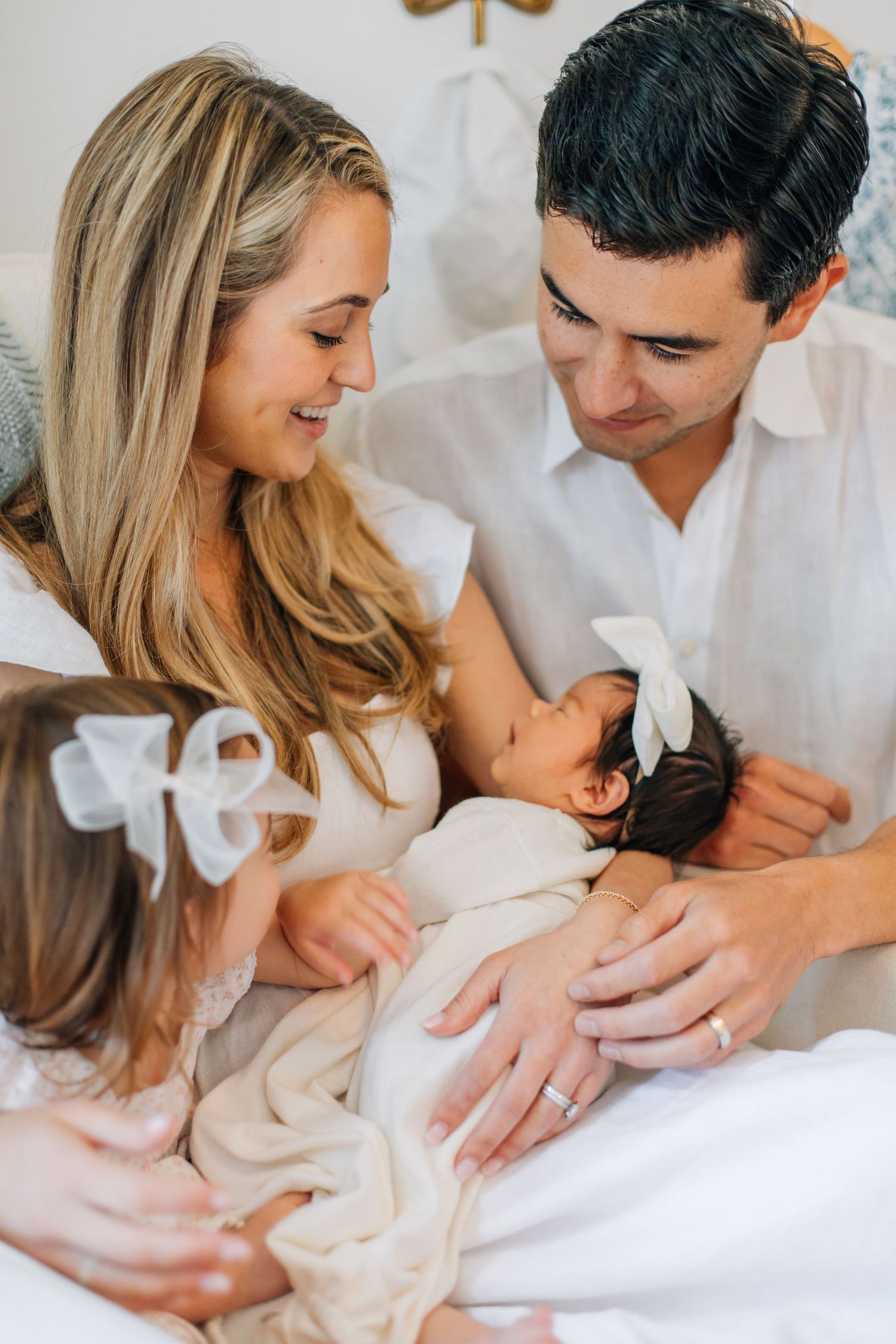 Loving family of four with blonde mother, dark-haired father, toddler, and newborn on white bed.