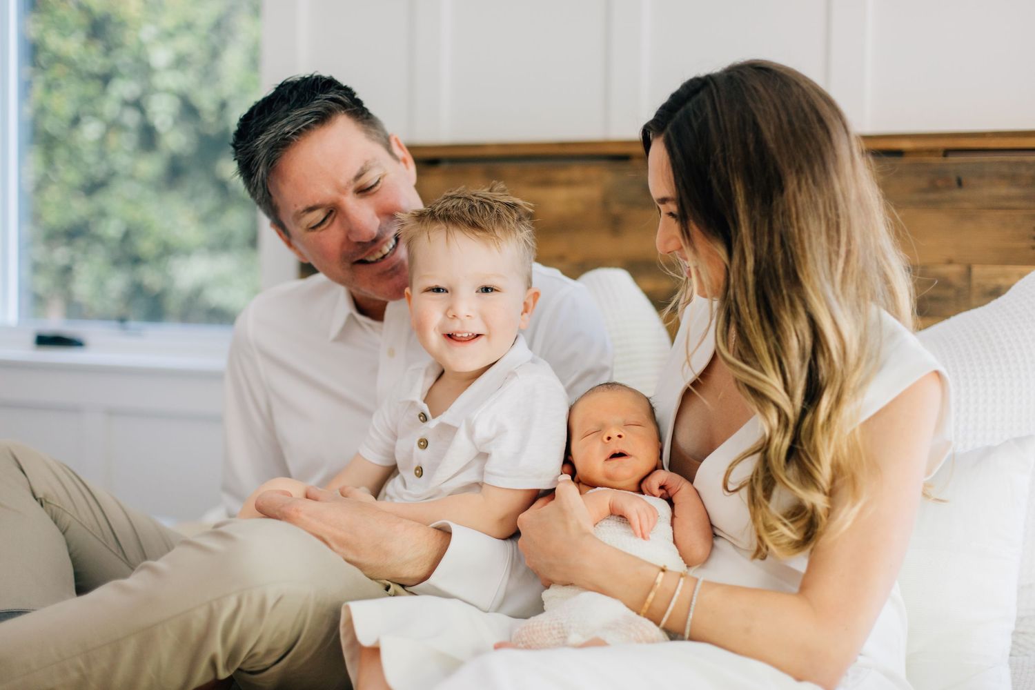 Joyful family with father, toddler son, and mother holding newborn baby on a white bed.