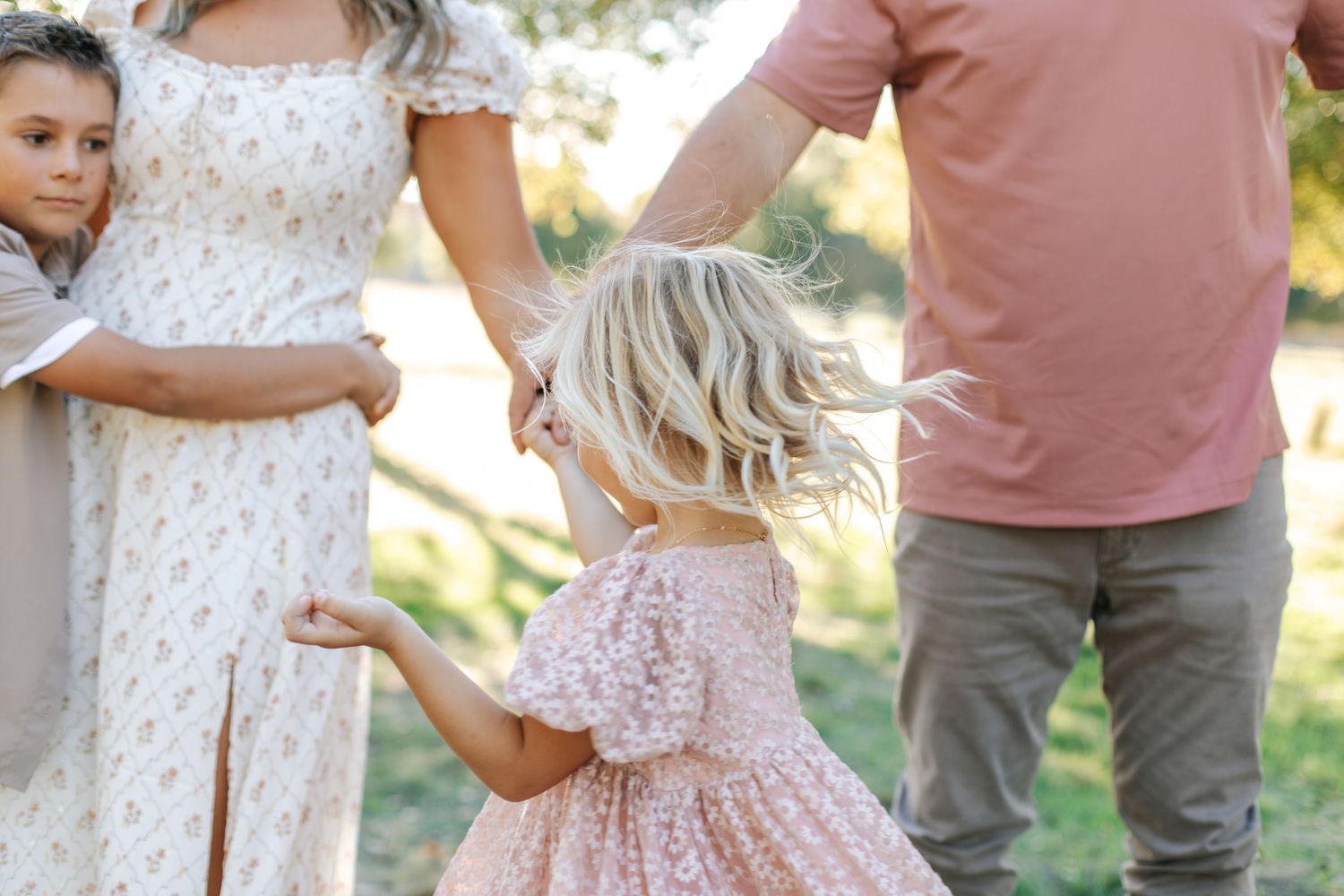 Young blonde toddler in pink floral dress spinning between parents in a sunlit outdoor setting.