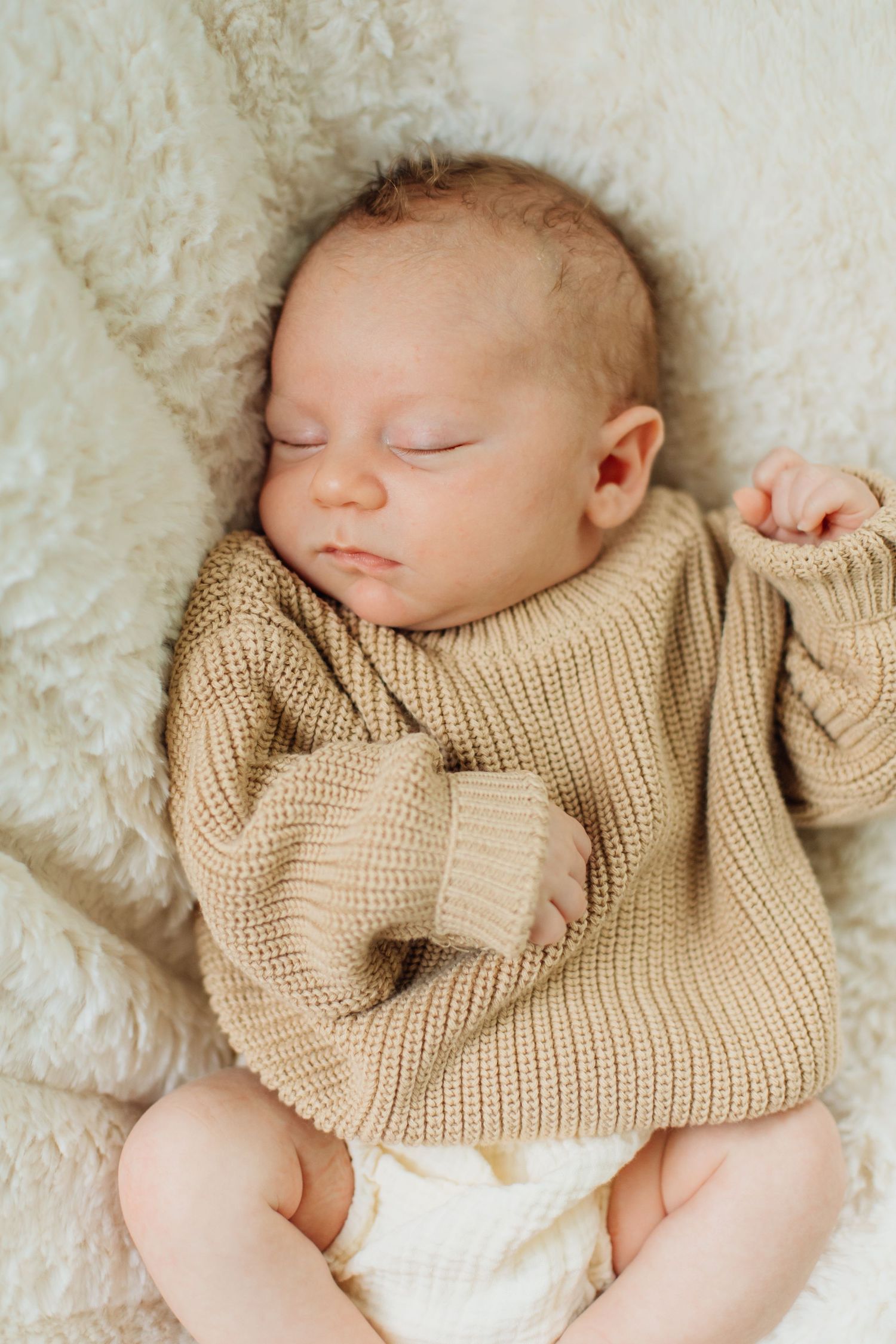 Peaceful newborn baby sleeping in oversized beige knit sweater on fluffy white blanket.