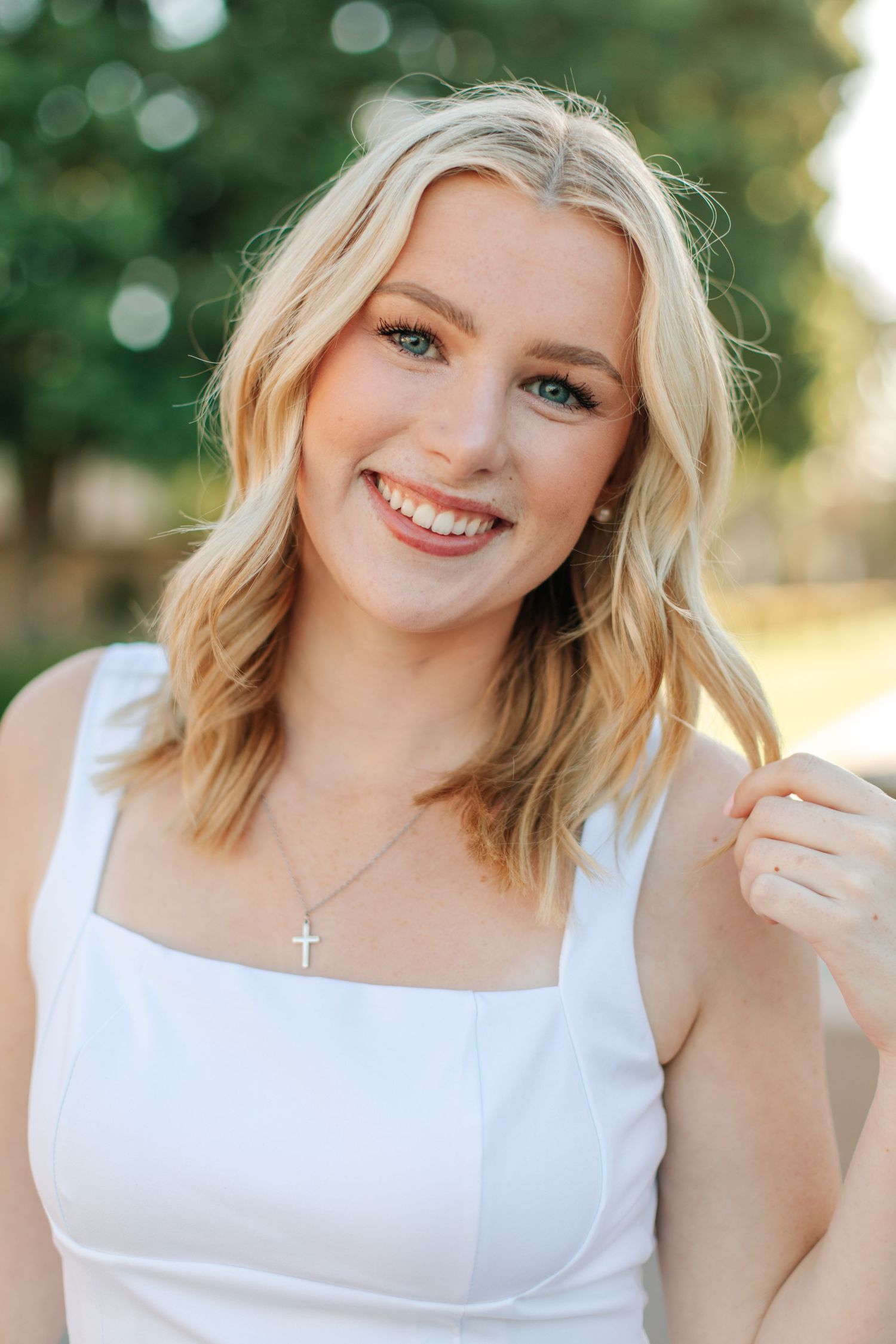 A smiling blonde young woman wearing a white top and a delicate cross necklace outdoors.