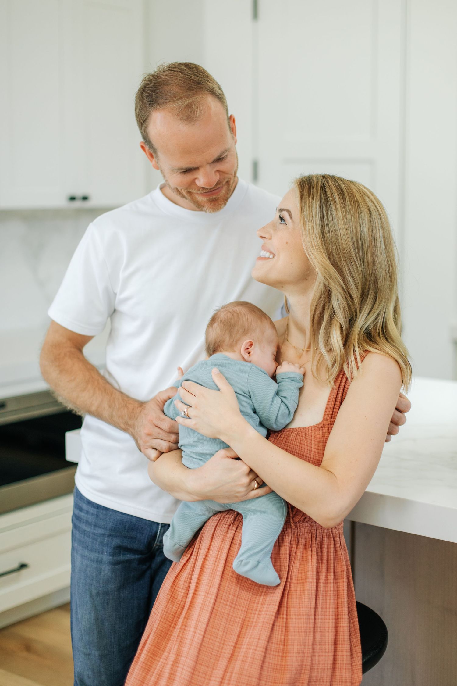 Happy couple smiling at each other while mother holds newborn baby in light blue onesie in modern white kitchen.