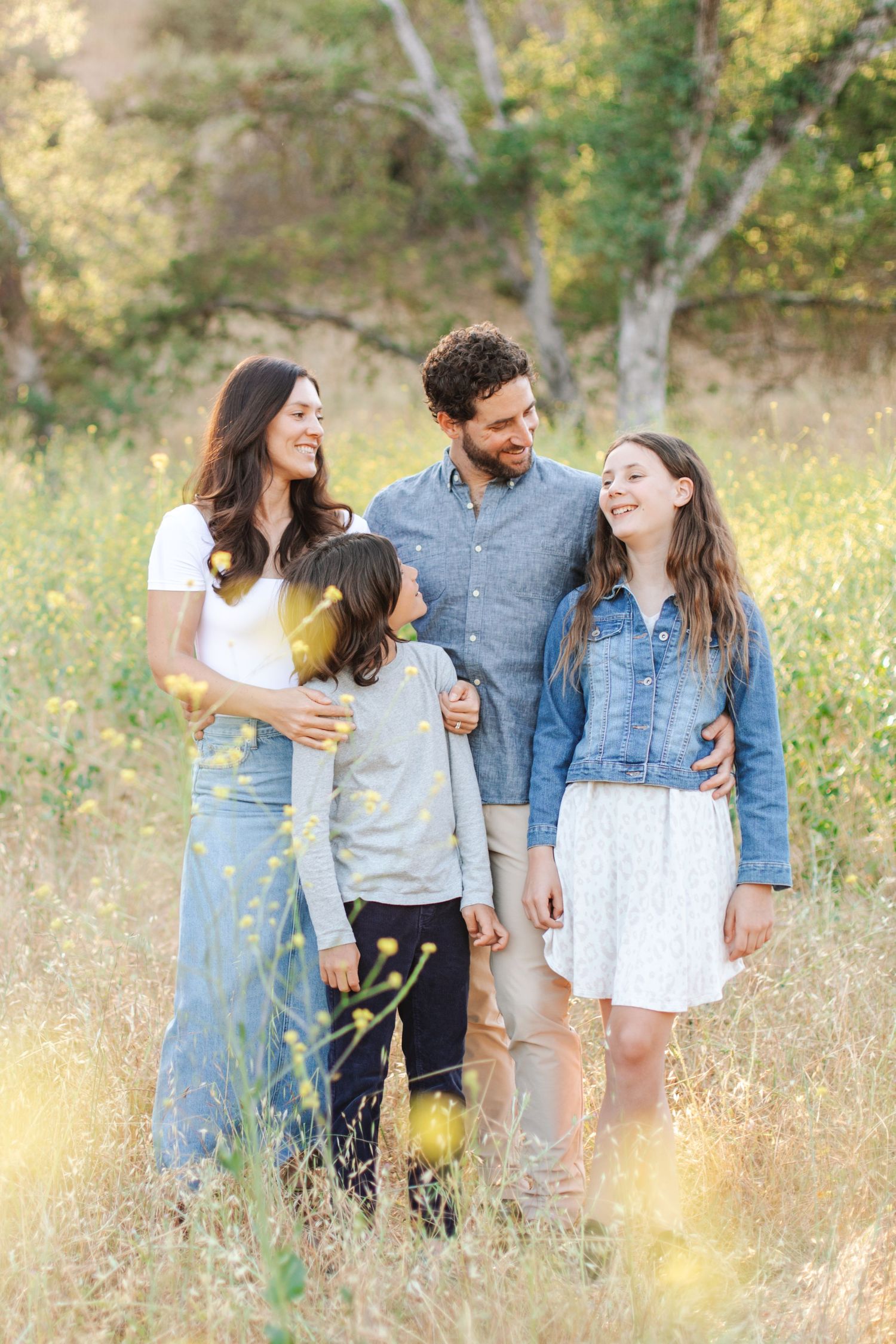 Happy family of four walking through wildflower meadow, parents embracing children in warm natural light.