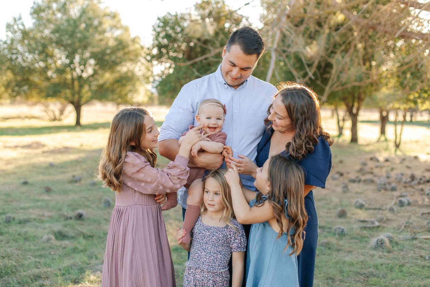 A happy family of six poses outdoors in a sunlit field, with parents holding a smiling baby surrounded by three young daughters.