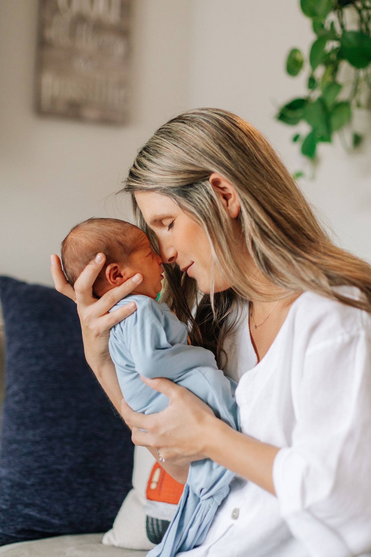Blonde mother in white shirt tenderly kissing her newborn baby wrapped in blue swaddle.