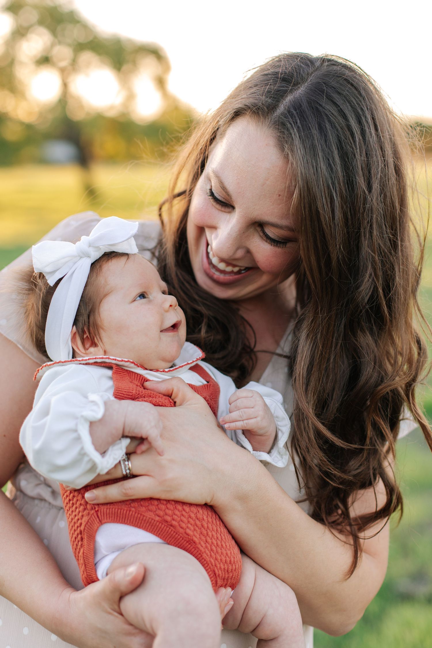 A smiling mother holds her newborn baby dressed in red and white outdoors in golden sunlight.