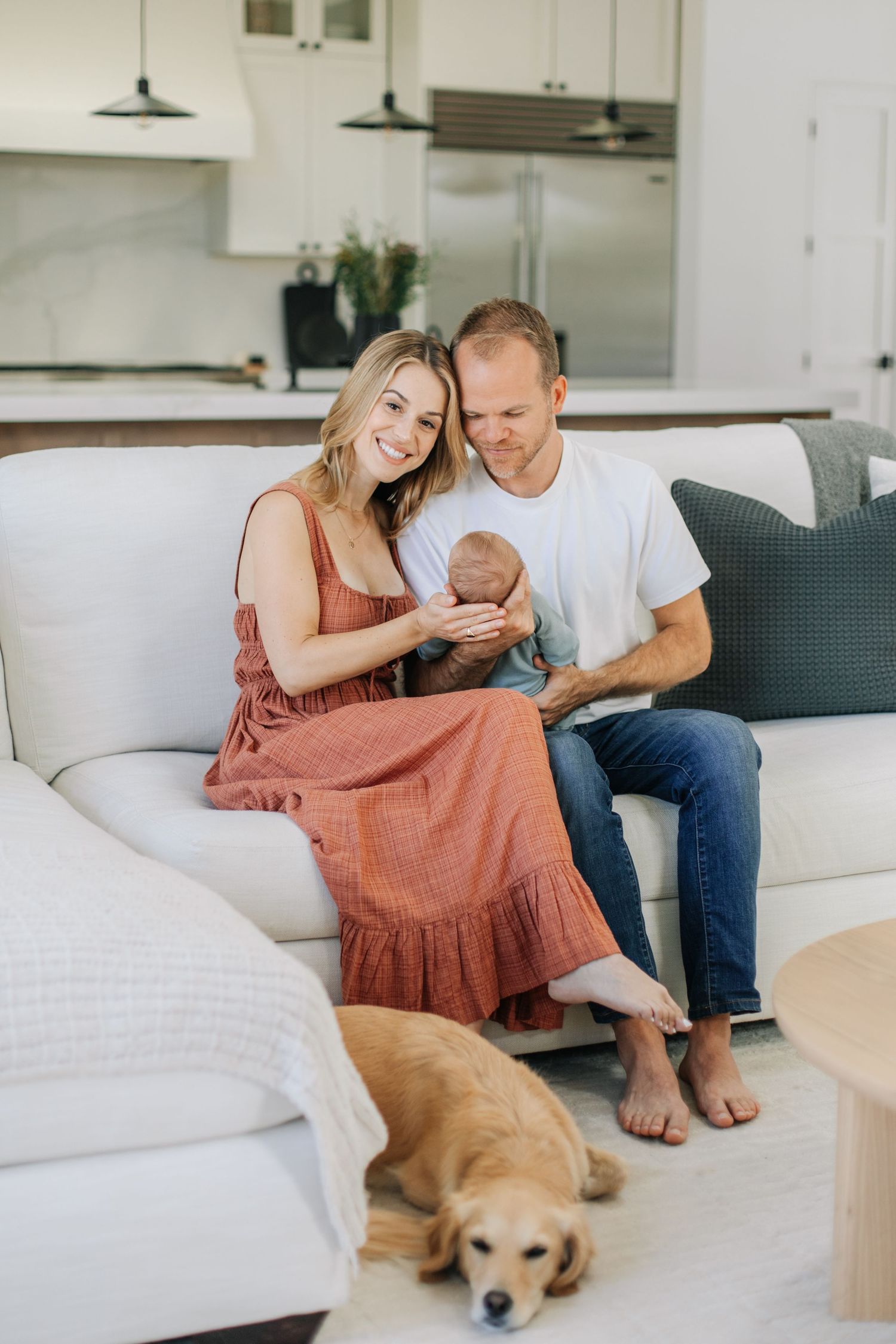 Smiling couple on a white sofa holding their newborn baby with a golden retriever resting at their feet.