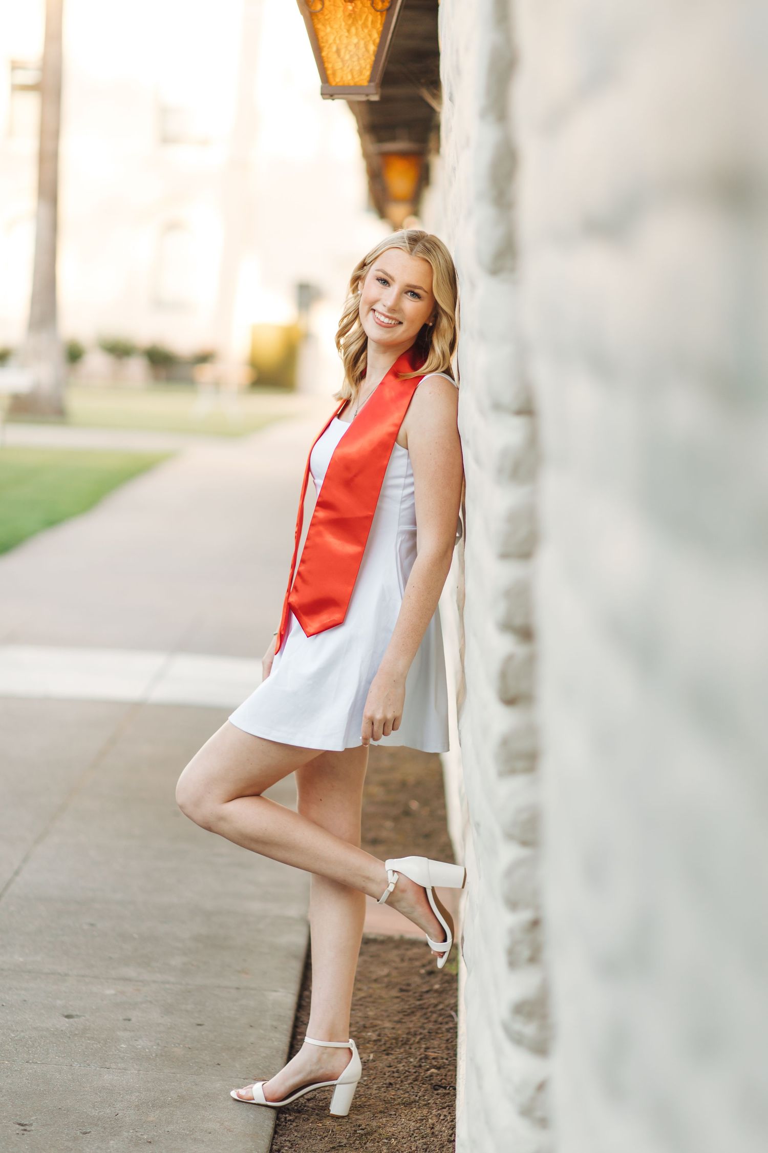 Smiling blonde graduate in white dress with red sash leaning against stone wall outdoors.