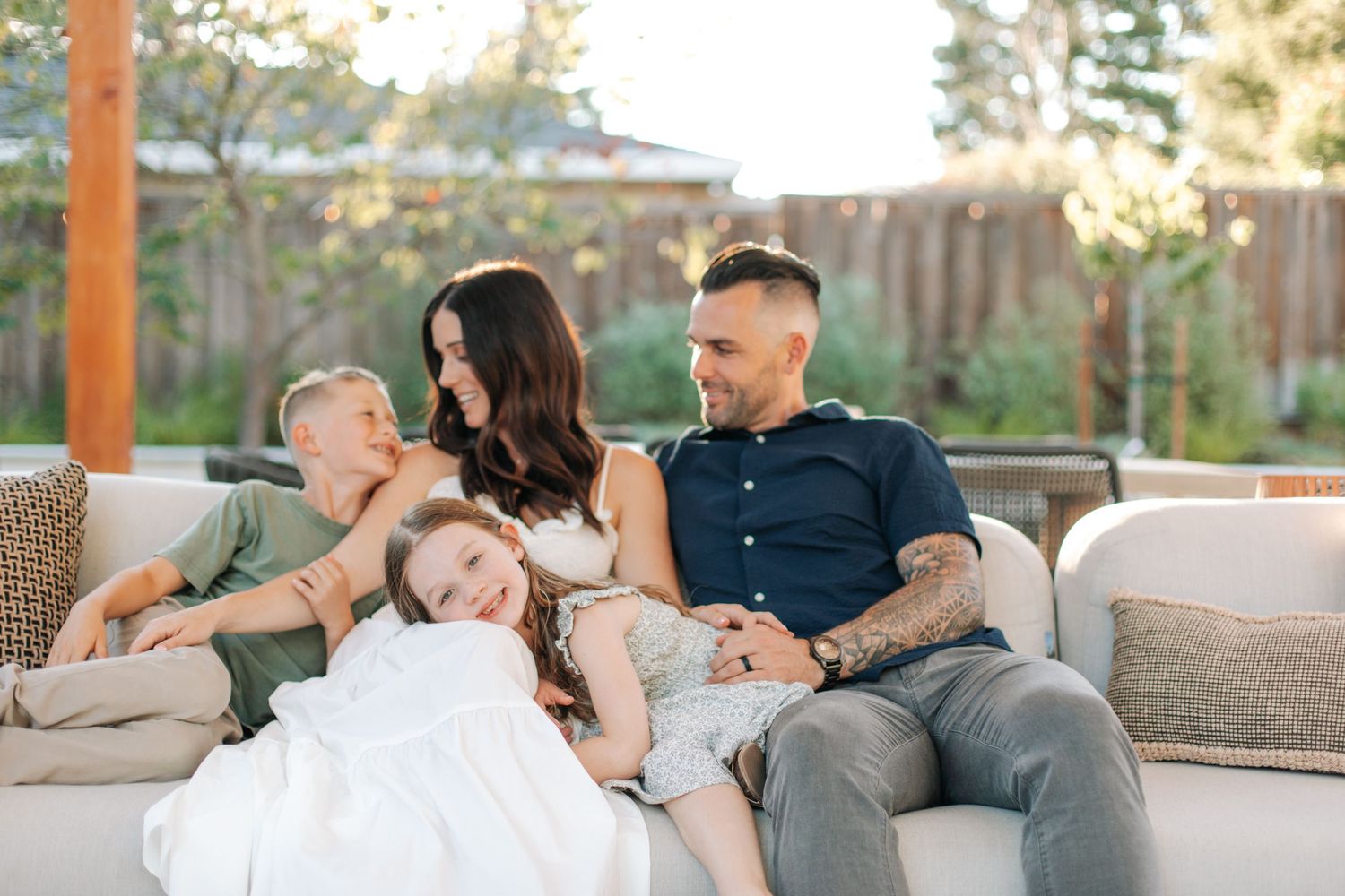 A happy family of four relaxes together on an outdoor patio sofa in a sunny backyard.