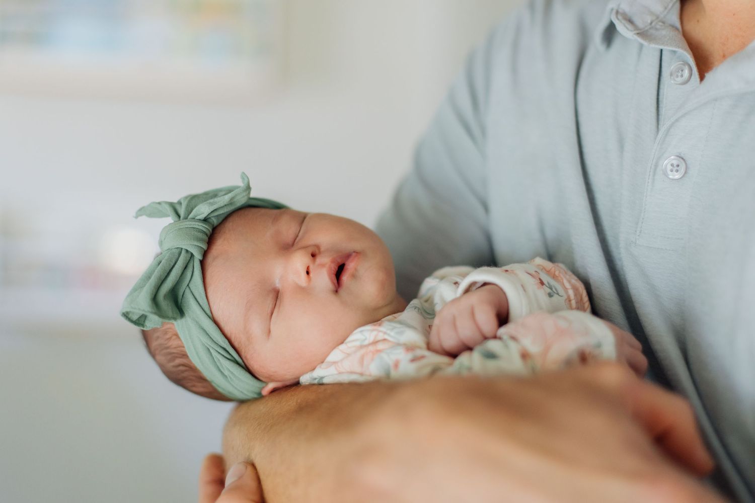 Newborn baby girl sleeping peacefully in fathers arms wearing a green bow headband and floral outfit.