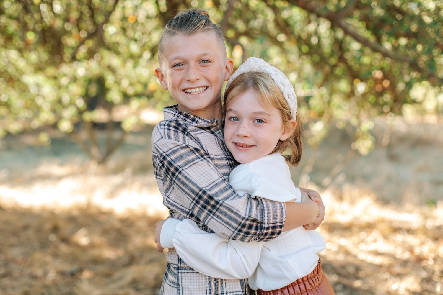 Two siblings hugging and smiling under a tree, older boy in plaid shirt embracing younger girl.