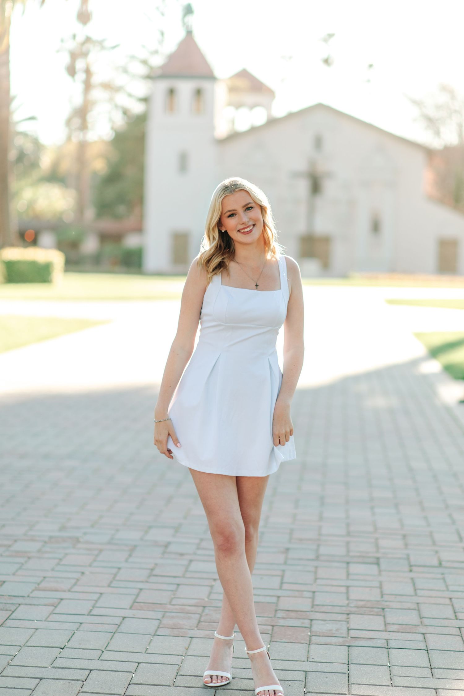 Young blonde woman in white mini dress and heels smiling on brick path with historic building behind her.