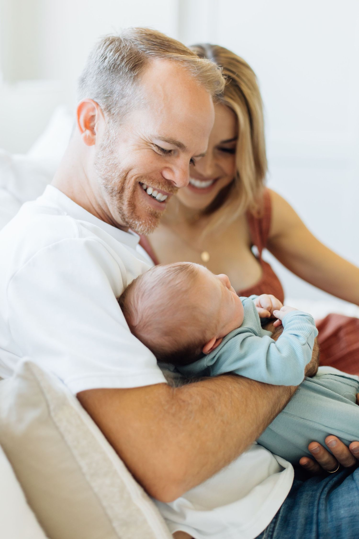 Laughing father holding newborn baby in blue onesie while smiling mother looks on from behind on sofa.
