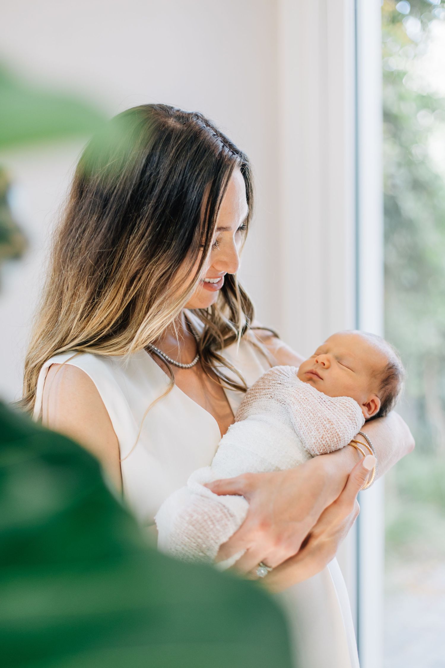 Loving mother in white dress holds her sleeping swaddled newborn baby by a bright window.