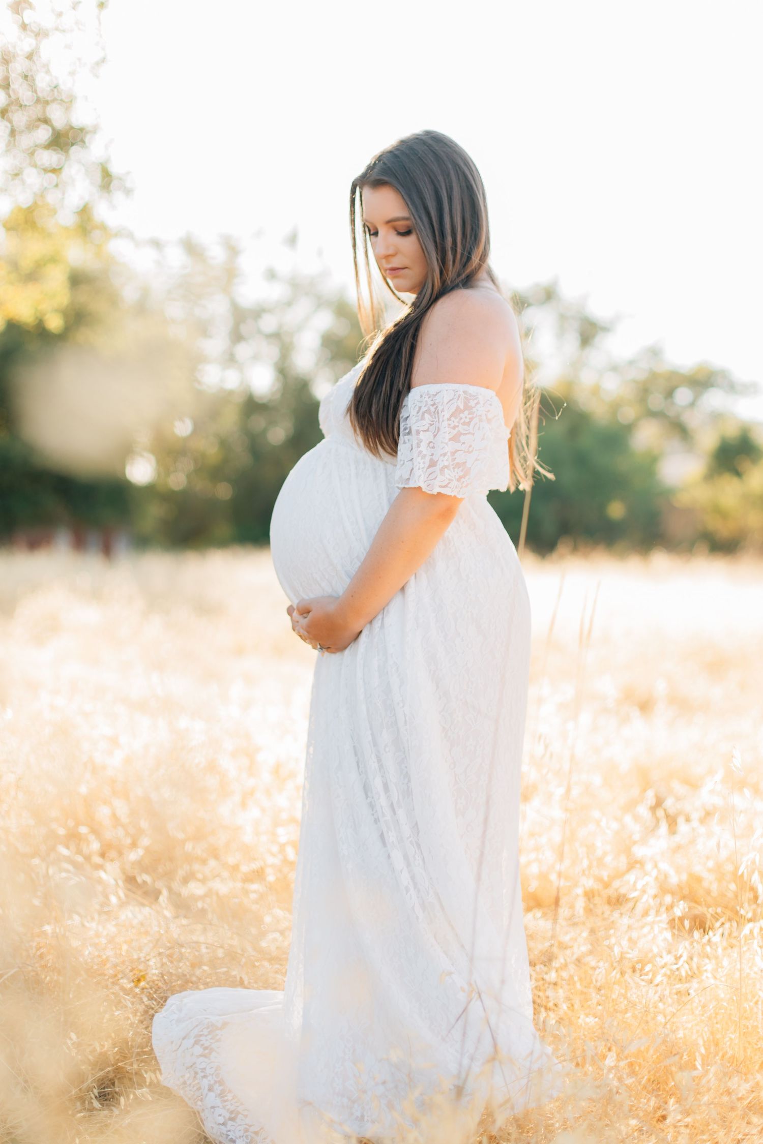 Pregnant woman in white lace off-shoulder gown cradling baby bump in golden field at sunset.