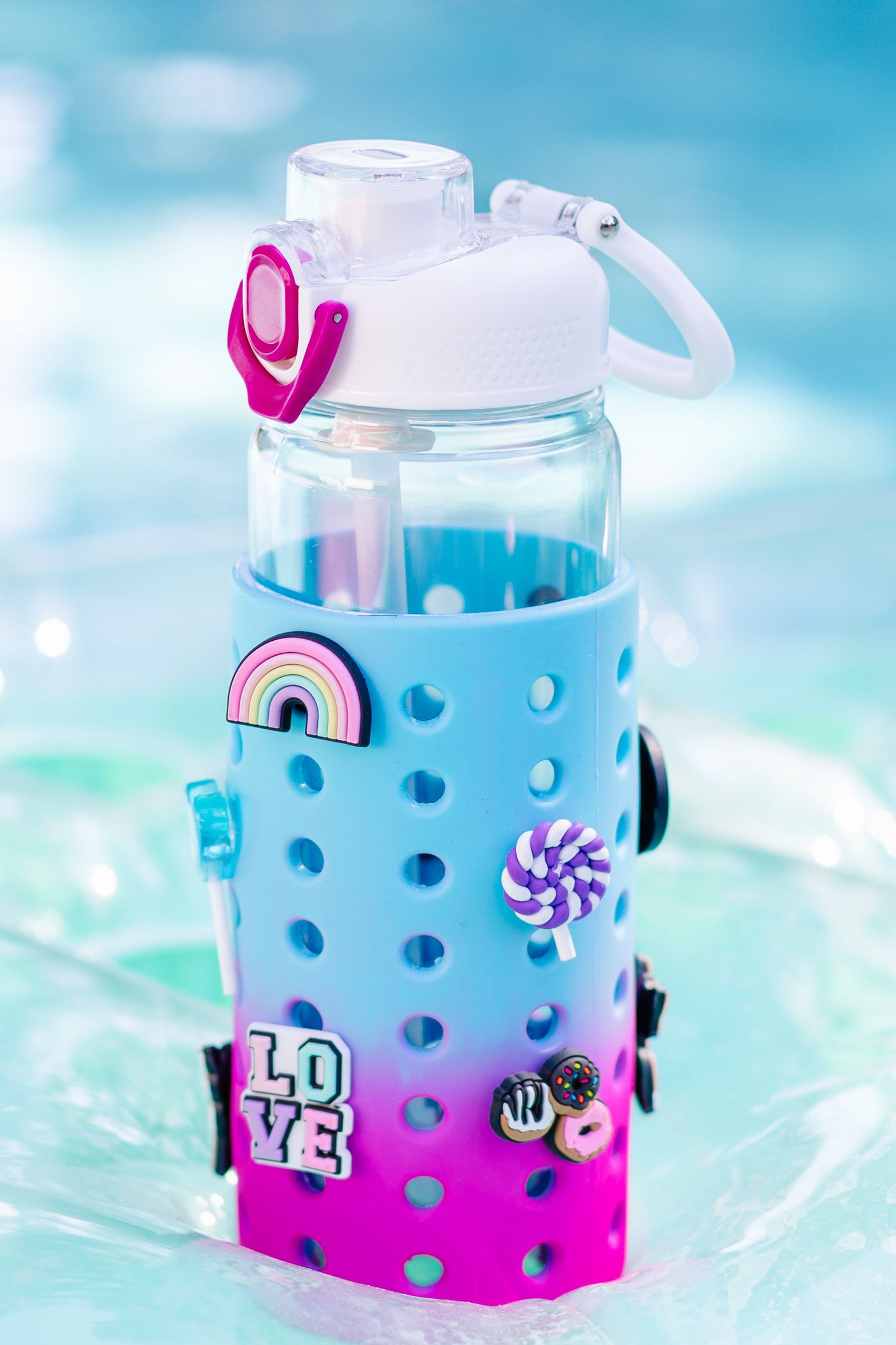 Blue and pink ombre silicone sleeve water bottle with rainbow and love charms by a pool.
