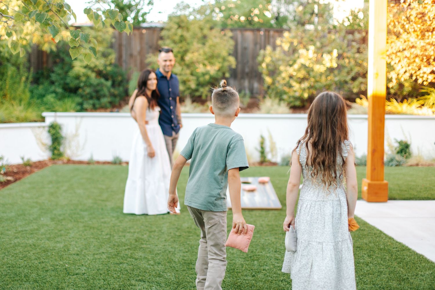 A family plays cornhole in a lush backyard, with two children tossing bean bags toward parents.