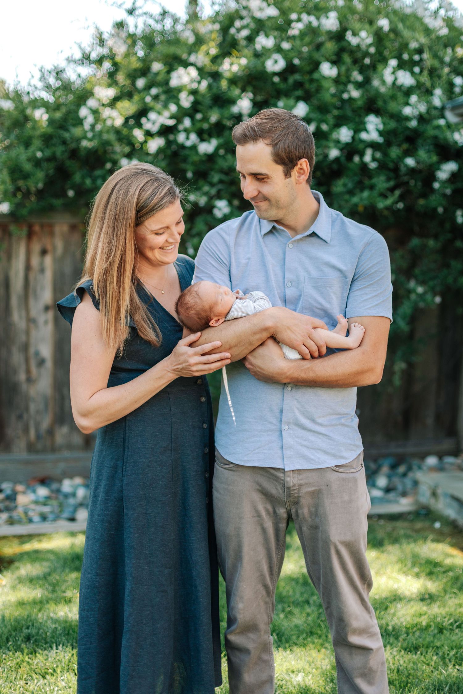 Happy couple standing in sunny backyard garden smiling down at newborn baby cradled in their arms.