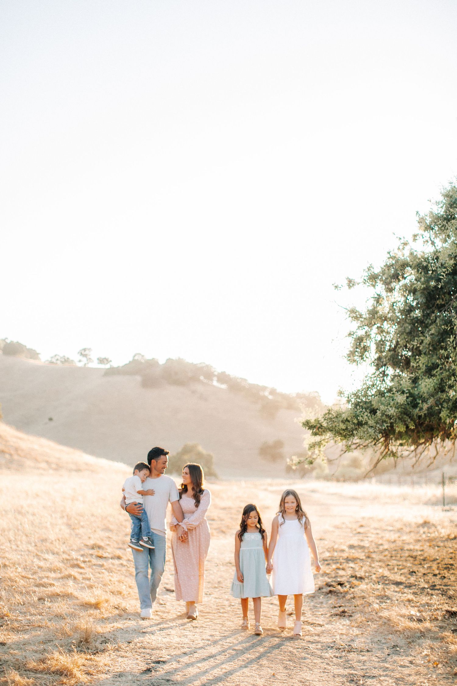 Family of five walking on a sunny dirt path through golden hills during a warm outdoor photo session.