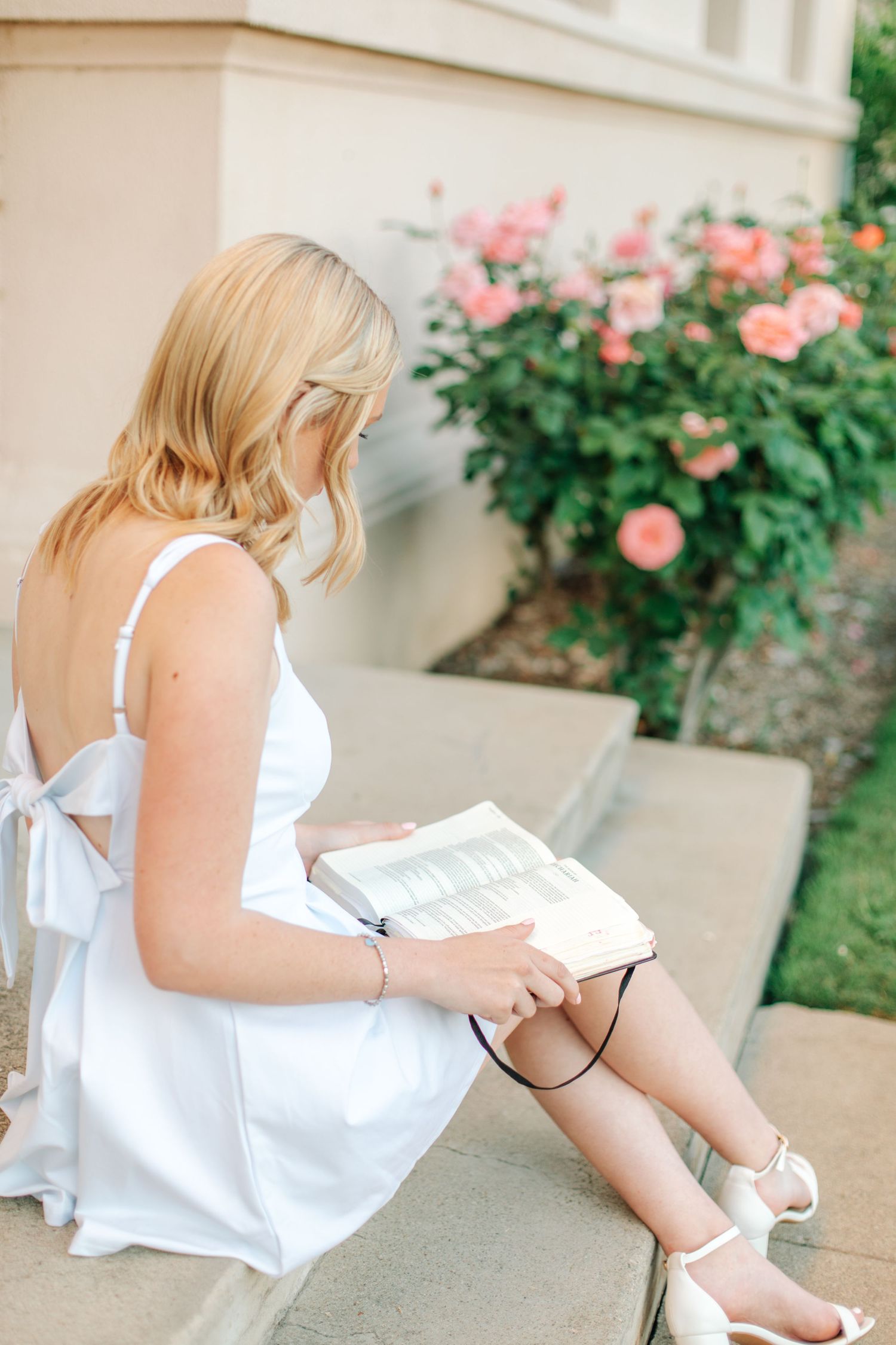 Blonde woman in white backless dress sitting on steps reading a book near pink roses.