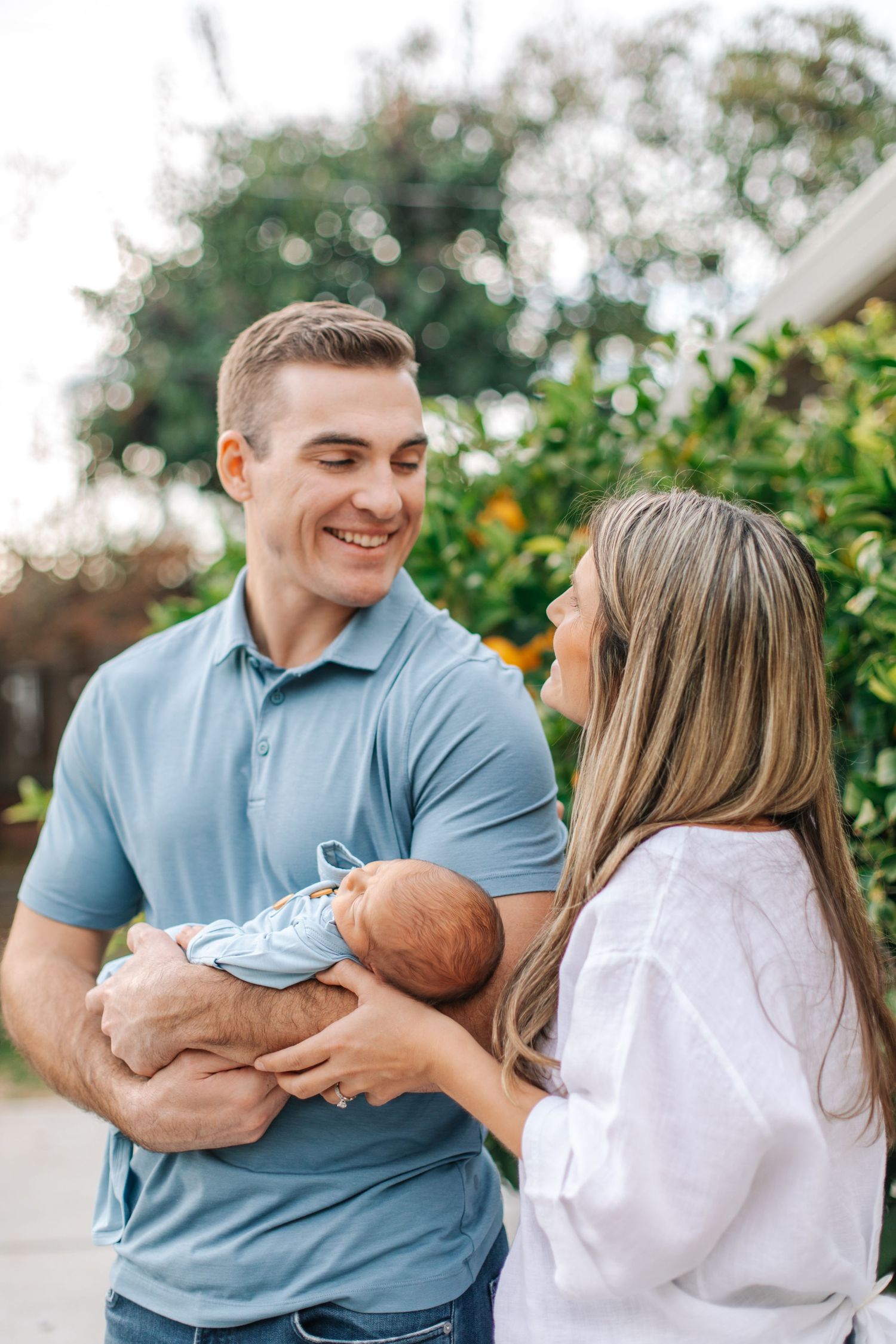 Smiling couple in blue and white outfits holding newborn baby together outdoors.