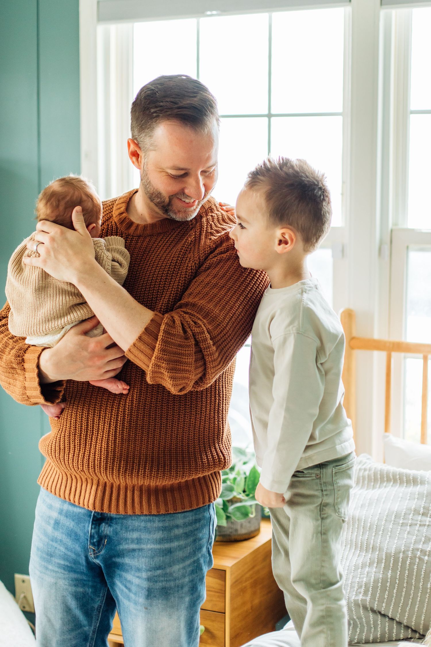 A smiling father in a brown sweater holds his newborn while his young son stands beside him.