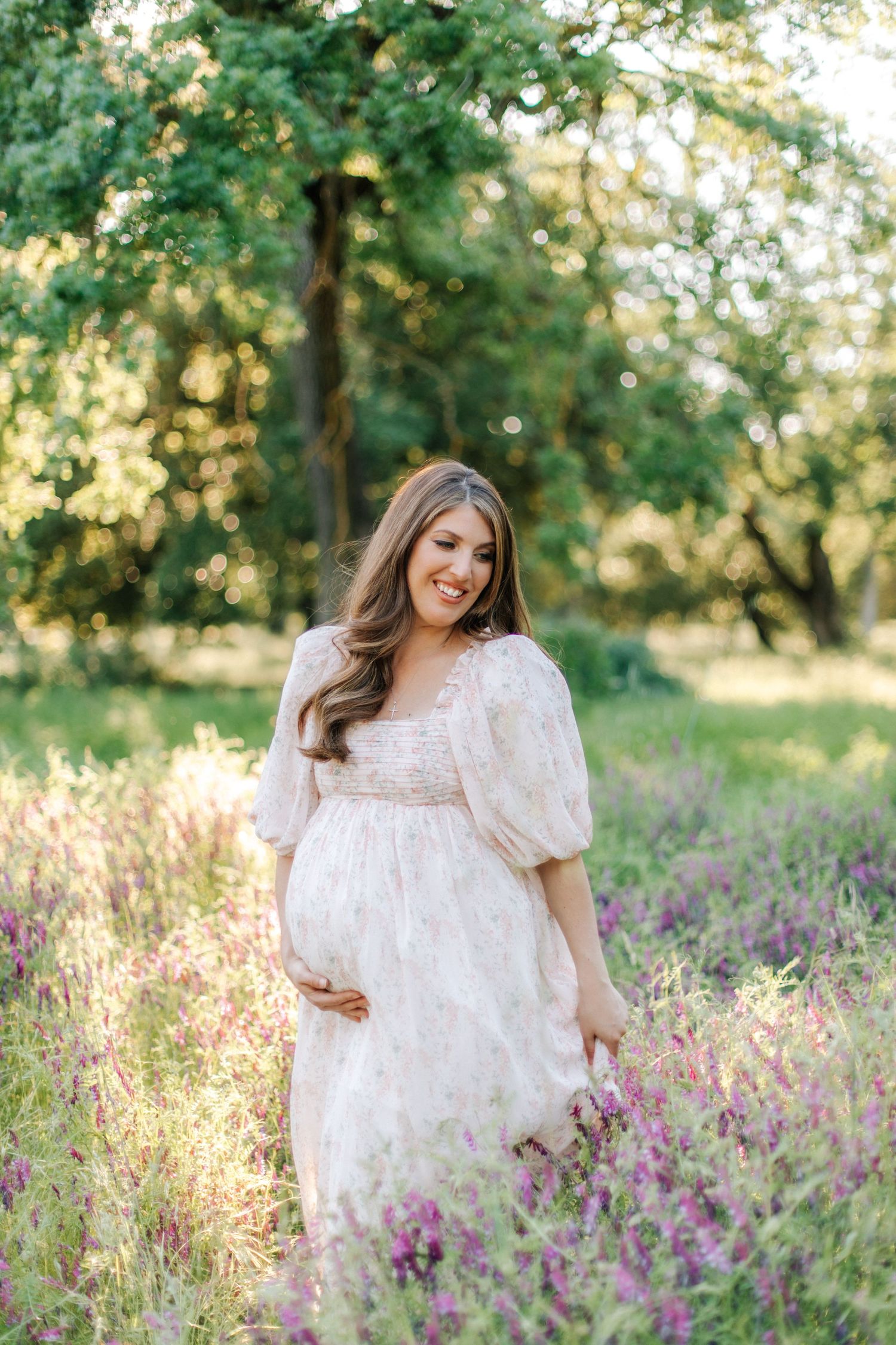 Smiling pregnant woman in white lace dress holding belly among purple wildflowers in sunlit garden.