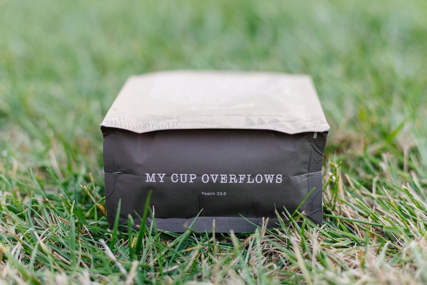 Brown kraft paper coffee bag with text My Cup Overflows sitting on green grass.