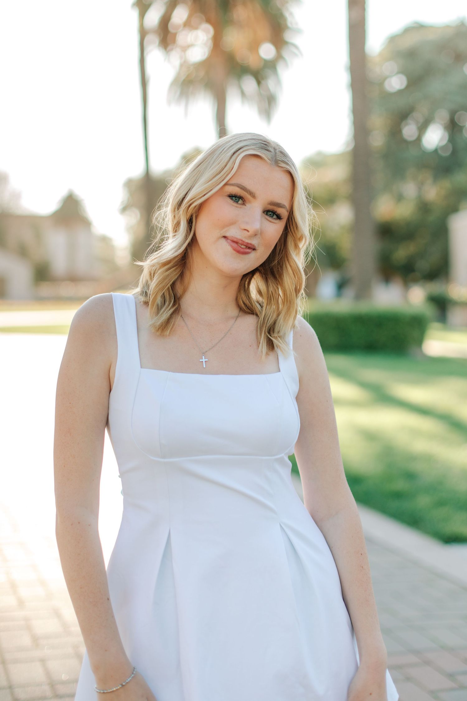 Smiling blonde woman in white sundress with cross necklace posing outdoors near green hedges and palm trees.
