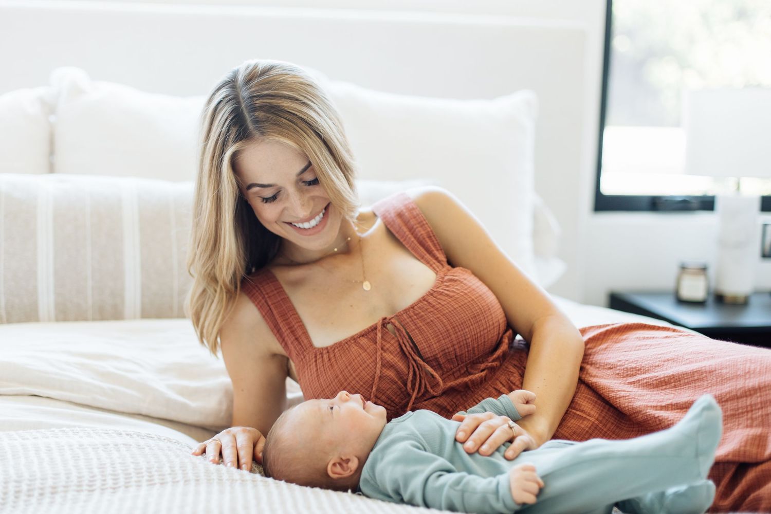 Smiling blonde mother in rust dress lying on white bed gazing lovingly at her newborn baby.