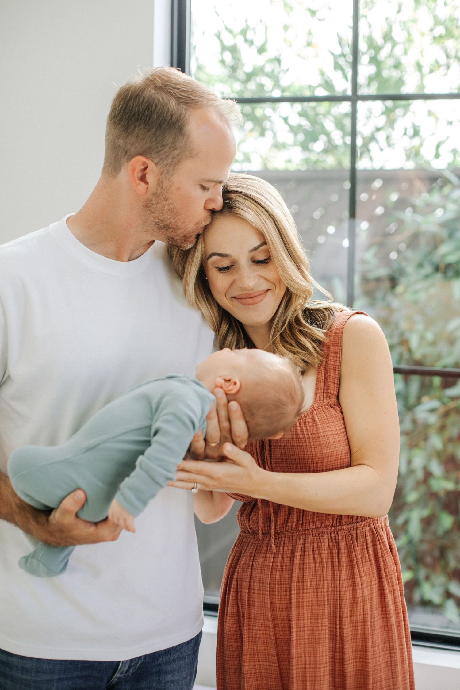 Father kissing mothers forehead as they both admire newborn baby in sage green sleeper.